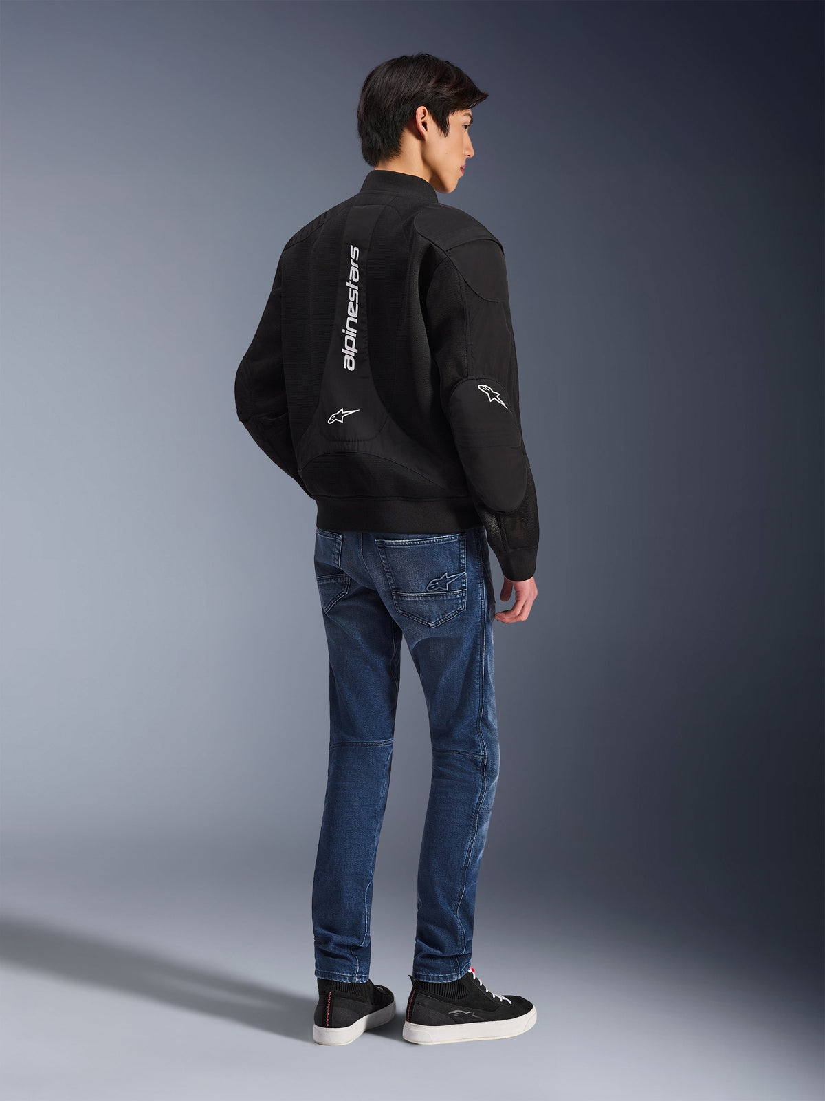 Modèle portant la veste Alpinestars Flight Air noire, vue de dos, associée à un jean de moto bleu et des baskets noires, avec marquage Alpinestars vertical blanc et panneaux de ventilation en maille 3D.