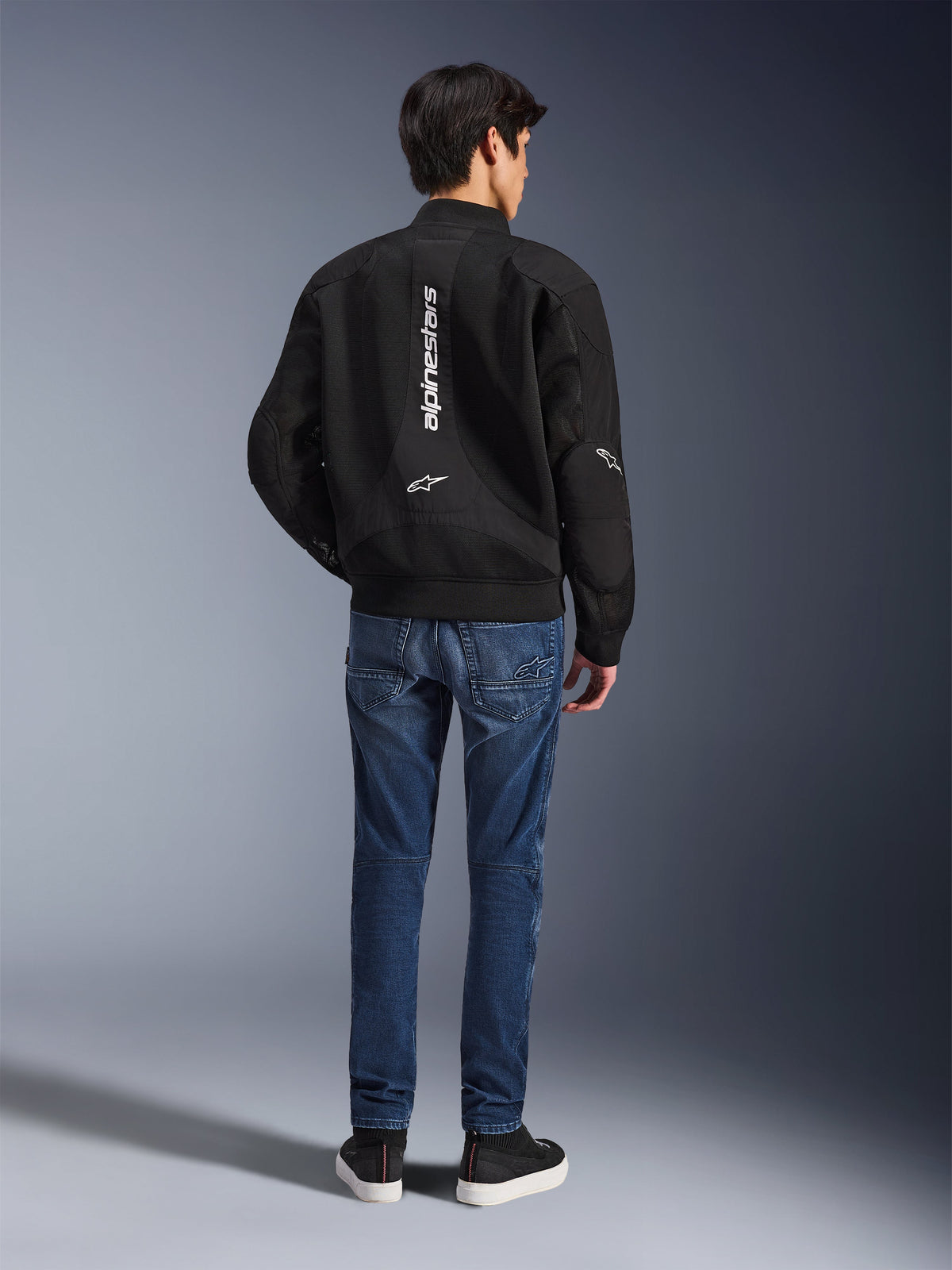 Modèle portant la veste Alpinestars Flight Air vue de dos, veste de moto noire style bomber avec panneaux en maille 3D et logo blanc vertical, associée à un jean de moto en denim bleu et des baskets noires, équipement de moto urbain contemporain