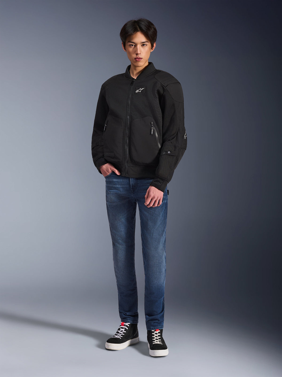 Modèle portant la veste Alpinestars Flight Air en noir, une veste de moto urbaine de style bomber avec des panneaux en maille 3D et une fermeture éclair frontale, associée à un jean bleu et des baskets noires, équipement de moto contemporain d'inspiration streetwear.