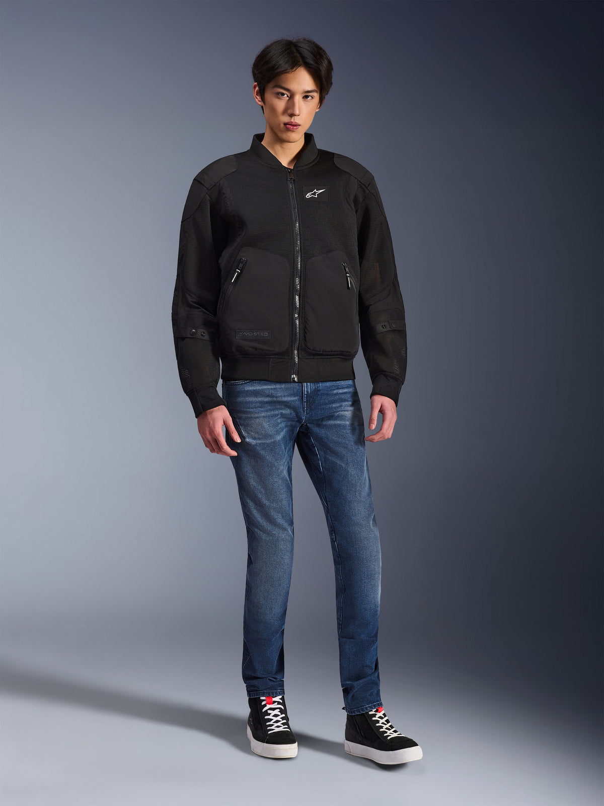 Veste Alpinestars Flight Air, noire, veste de moto de style bomber d'inspiration urbaine avec panneaux en maille 3D et logo blanc sur la poitrine, portée par un mannequin avec un jean bleu et des baskets noires, tenue de moto contemporaine
