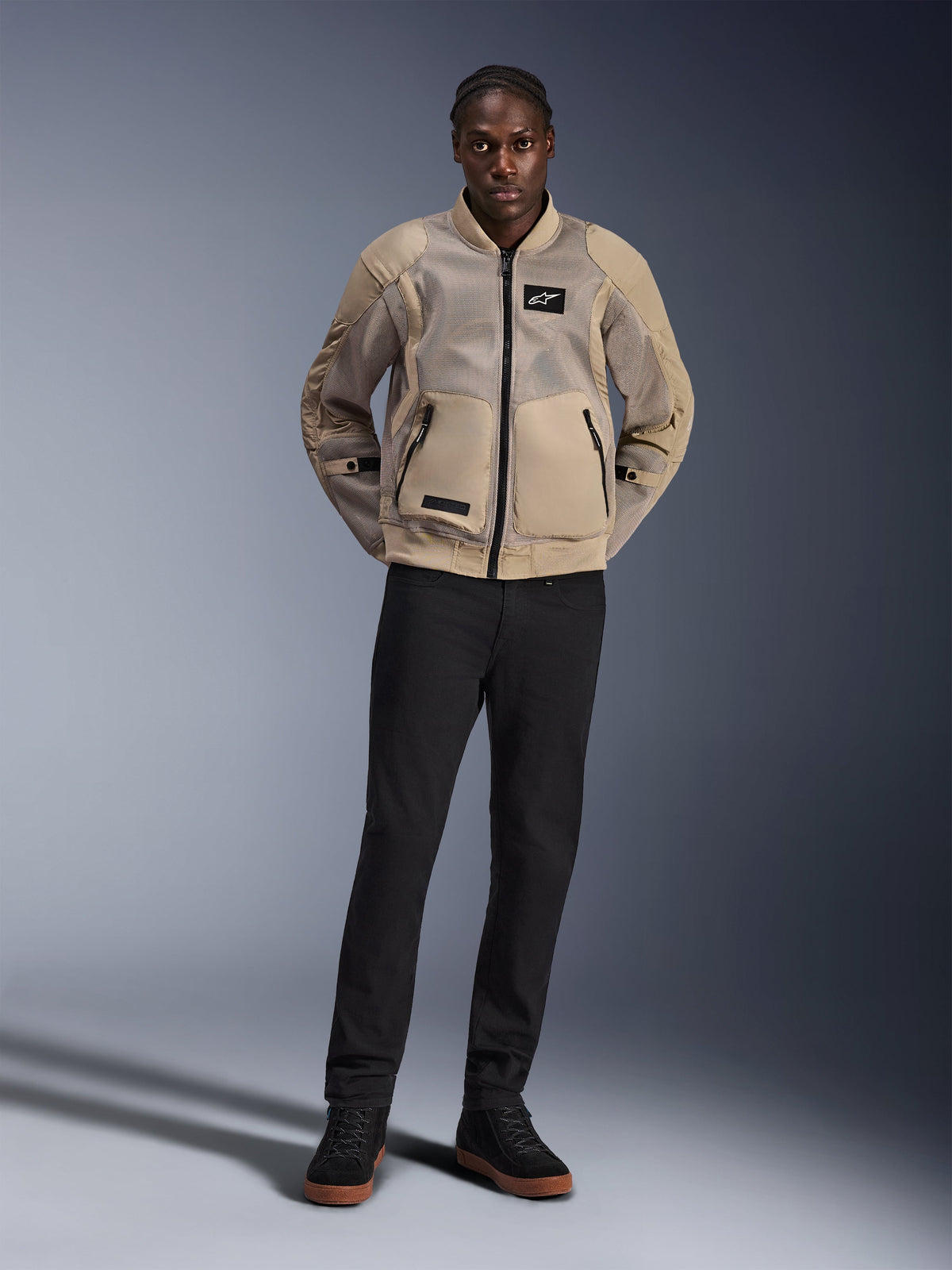 Mannequin portant la veste Alpinestars Flight Air en marron, veste de moto urbaine avec larges panneaux en maille 3D, col et poignets côtelés inspirés du style bomber, associée à un pantalon noir et des chaussures de moto noires, équipement urbain contemporain