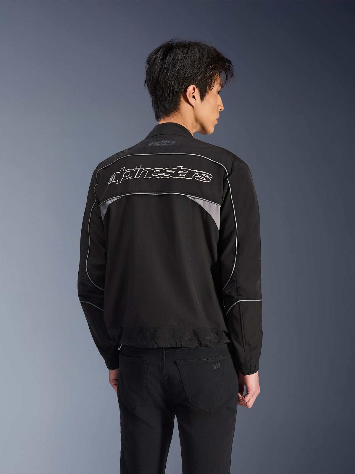 Veste Alpinestars Unite, veste de moto urbaine, noire avec des accents gris foncé, vue arrière montrant le lettrage Alpinestars blanc sur les épaules, liserés et panneaux réfléchissants, design streetwear minimaliste en softshell