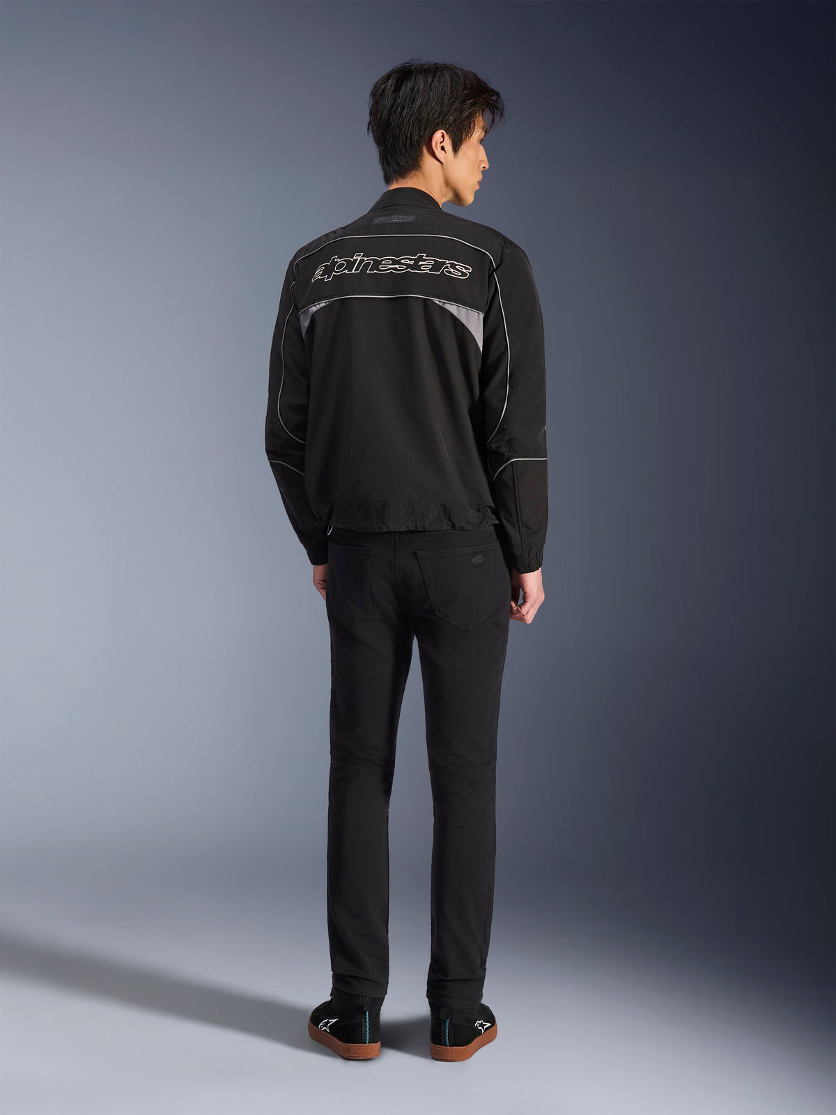 Veste Alpinestars Unite, veste de moto noire et gris foncé, vue arrière montrant le logo Alpinestars sur le dos, panneaux réfléchissants gris, design urbain minimaliste, portée avec un pantalon noir et des chaussures noires