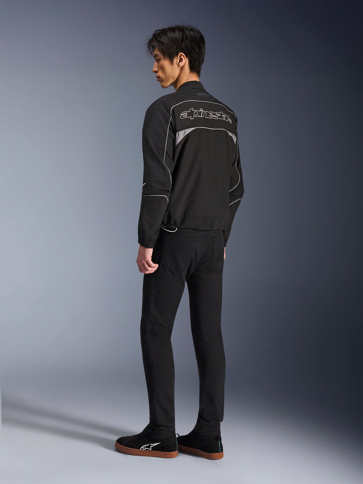 Veste Alpinestars Unite, veste de moto urbaine, noir et gris foncé, vue de dos montrant un grand logo Alpinestars à contours, lisérés et empiècements réfléchissants, design softshell minimaliste, portée avec un pantalon noir et des chaussures décontractées