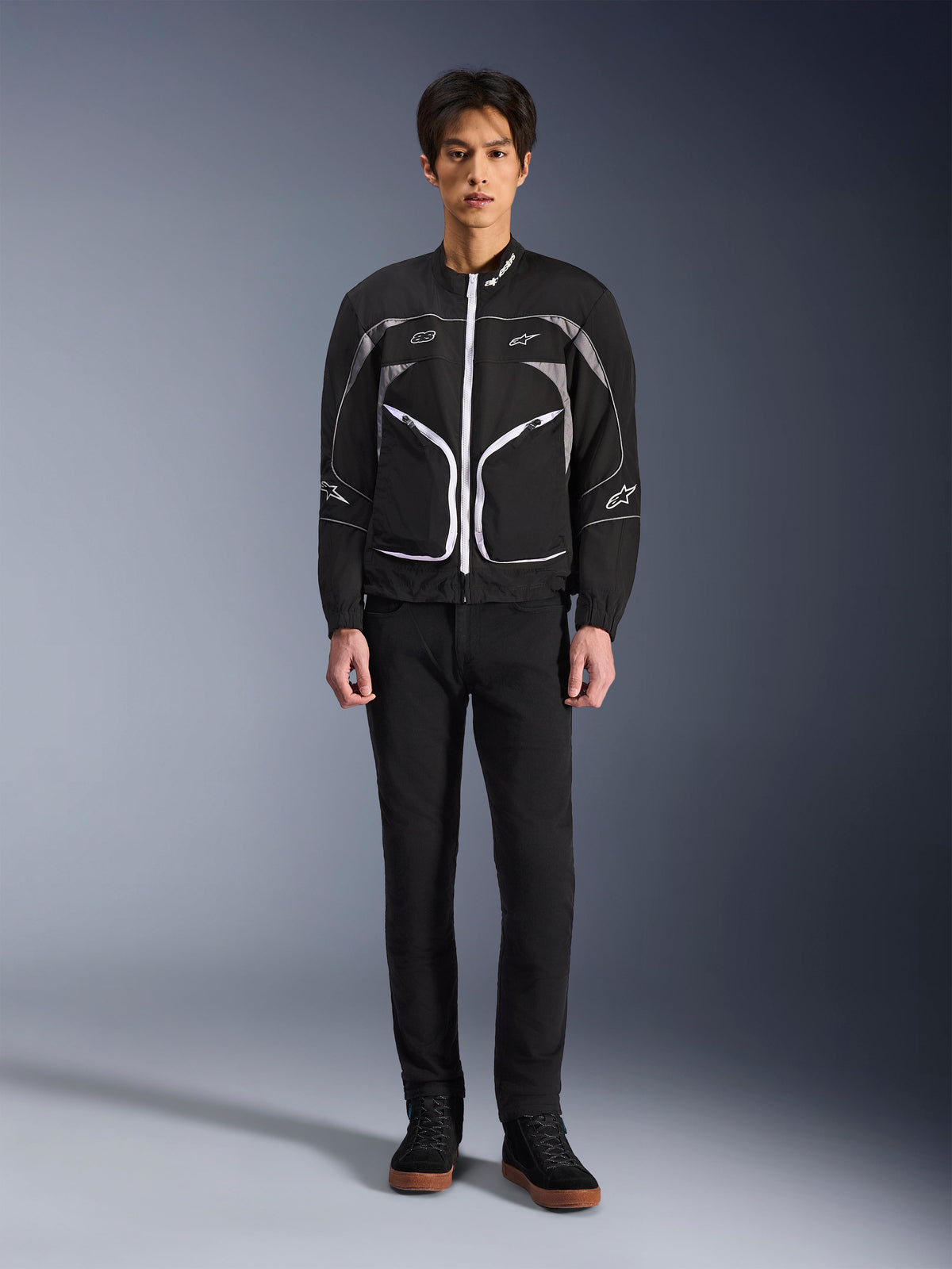 Veste Alpinestars Unite, veste de moto, noir et gris foncé, portée par un mannequin avec un pantalon noir et des bottes noires, dotée de fermetures éclair contrastées blanches, de liserés réfléchissants et d'un design urbain minimaliste
