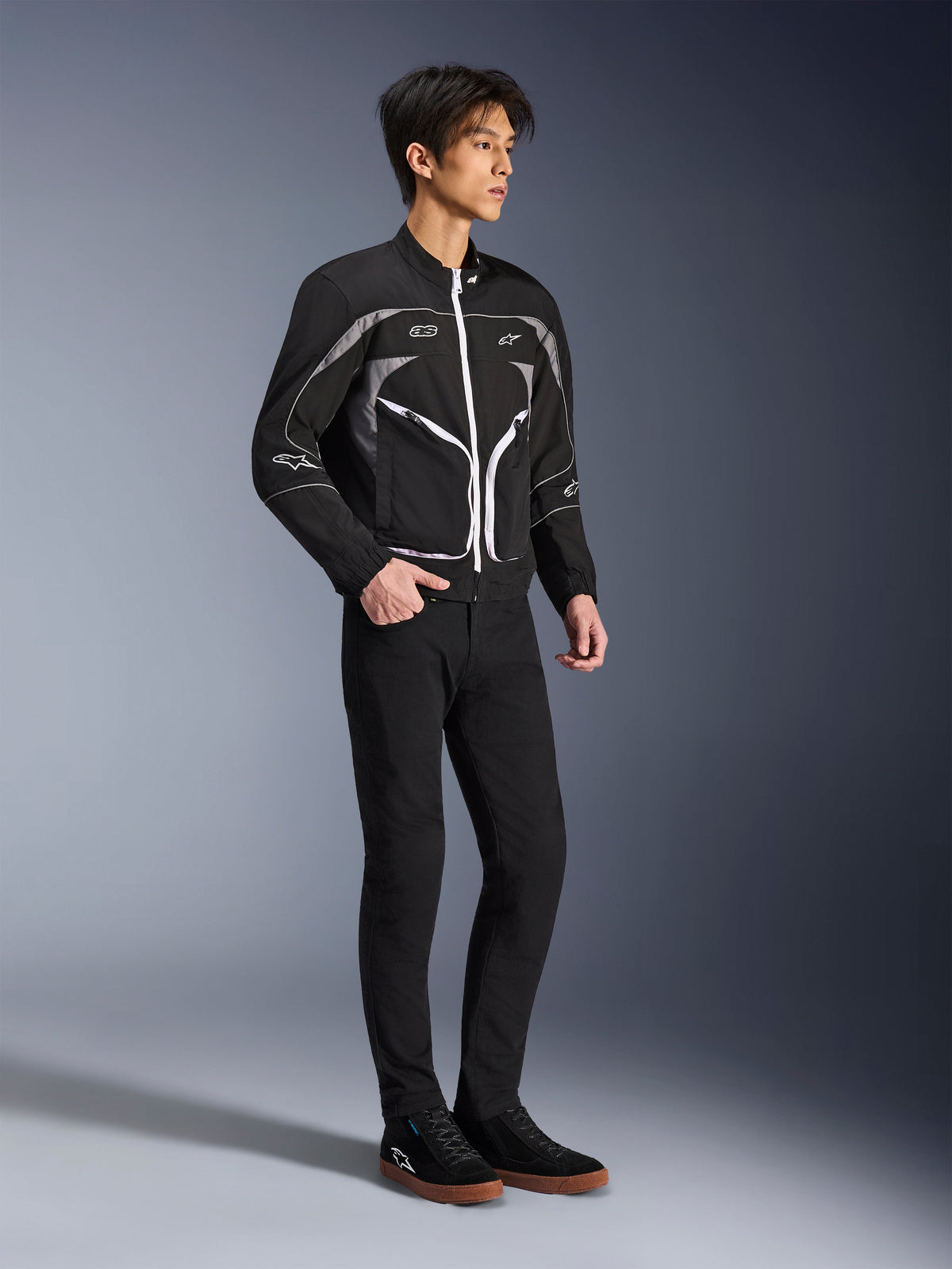 Mannequin portant la veste Alpinestars Unite en noir et gris foncé, veste de moto urbaine avec fermetures éclair blanches et détails réfléchissants, associée à un pantalon noir ajusté et des baskets, équipement de moto minimaliste d'inspiration streetwear
