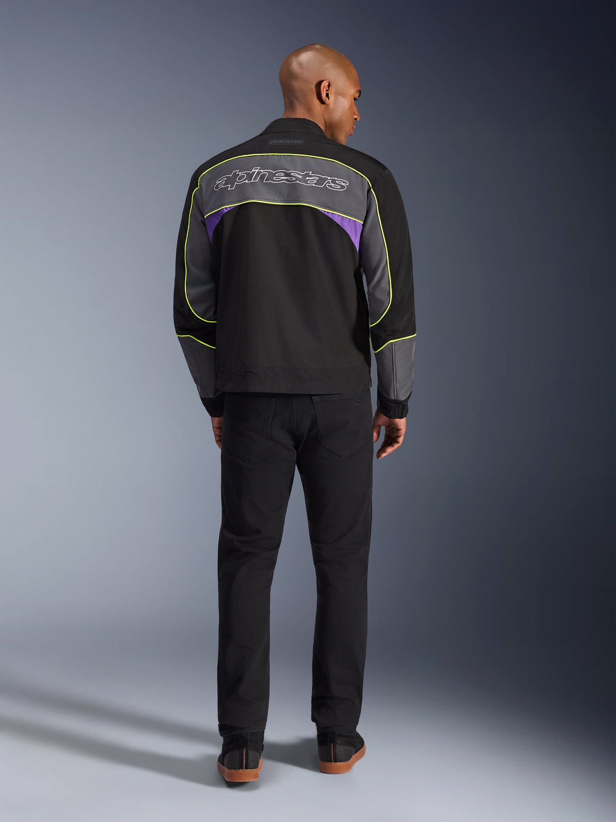 Veste Alpinestars Unite, veste de moto urbaine en Black Asphalt Violet, vue arrière montrant des empiècements gris avec des accents violets et un liseré jaune fluo, logo Alpinestars sur les épaules, portée avec un pantalon noir
