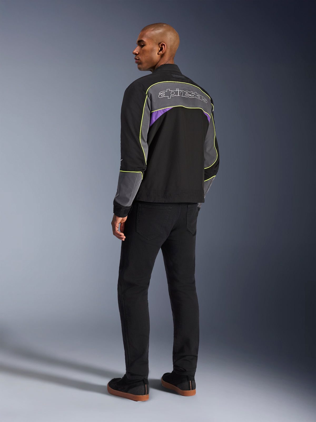 Veste Alpinestars Unite, veste de moto urbaine, Black Asphalt Violet, vue de trois-quarts arrière sur mannequin, softshell noir avec empiècements gris et accents violets, liseré réfléchissant jaune fluo, logo Alpinestars sur le haut du dos, associée à un pantalon noir et des chaussures décontractées noires