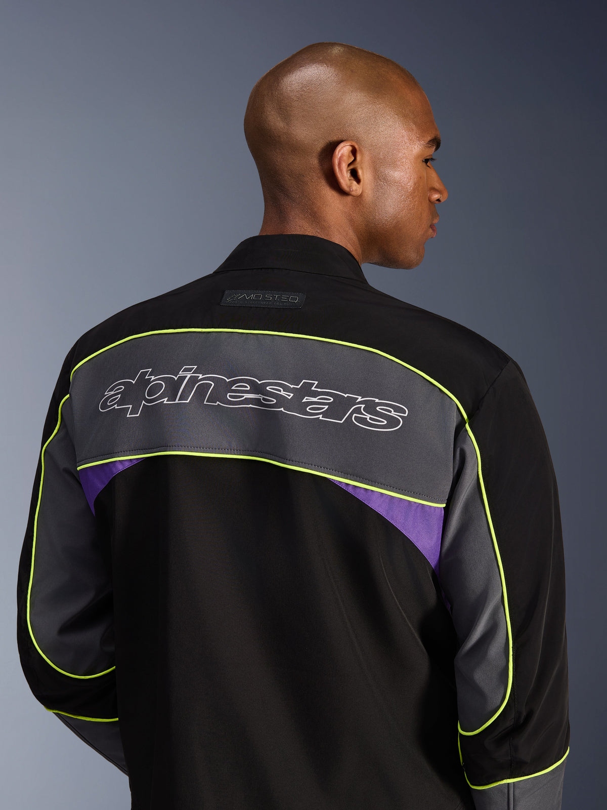 Veste Alpinestars Unite, veste de moto urbaine, Black Asphalt Violet, vue arrière montrant un large empiècement gris aux épaules avec logo Alpinestars, liseré jaune néon et accents violets sur une construction softshell à 3 couches