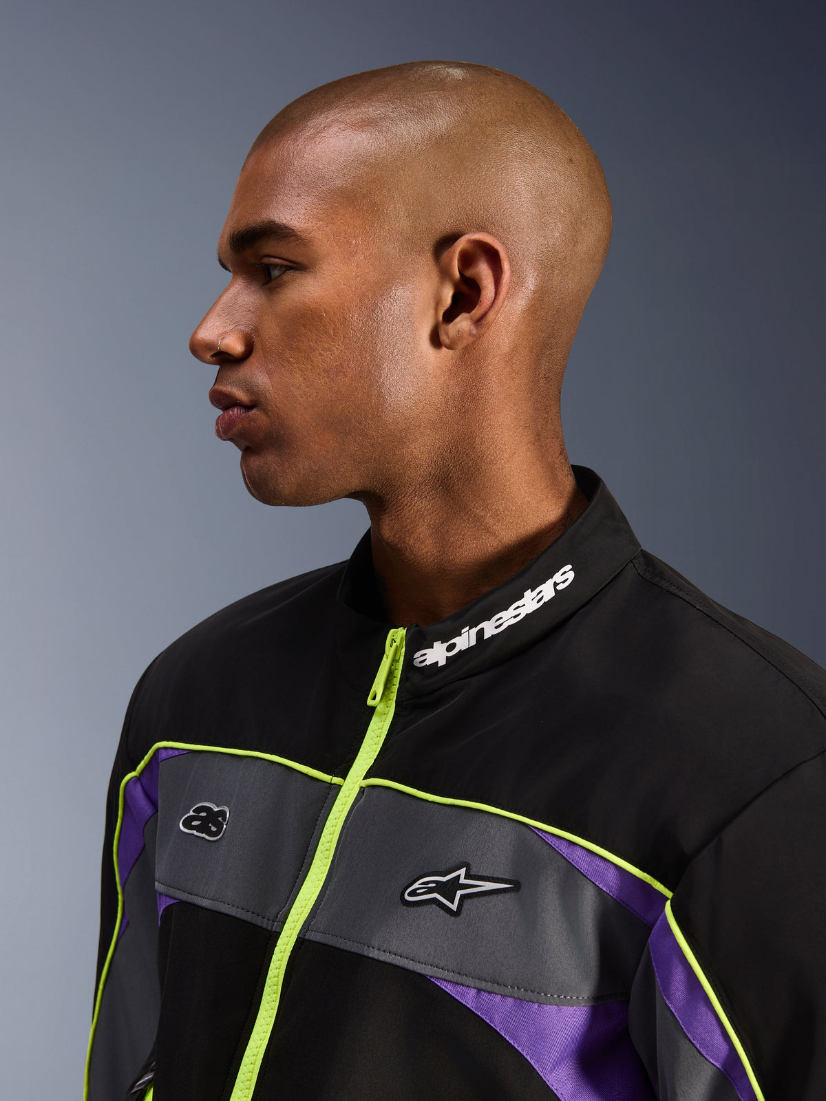 Veste Alpinestars Unite, Noir Asphalte Violet, vue de profil d'un mannequin homme centrée sur le col montant avec marquage Alpinestars blanc, fermeture éclair jaune fluo, empiècements gris sur la poitrine avec logos, accents violets, style streetwear moto urbain minimaliste