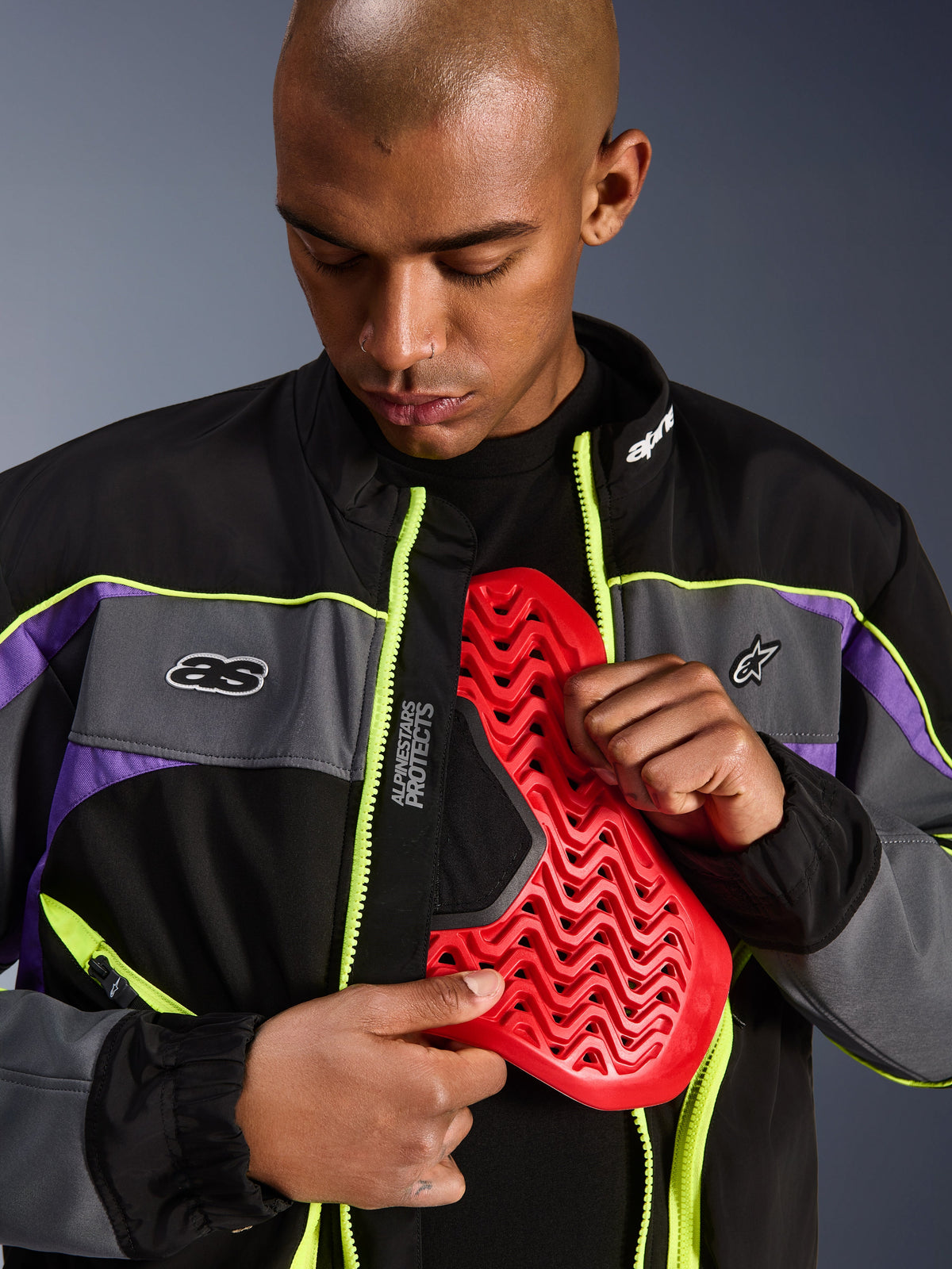 Veste Alpinestars Unite, veste de moto urbaine, noir asphalte et violet avec des accents jaune fluo, mannequin masculin présentant un insert de protection de poitrine Nucleon rouge à l'intérieur de la veste ouverte, esthétique streetwear minimaliste pour la conduite urbaine