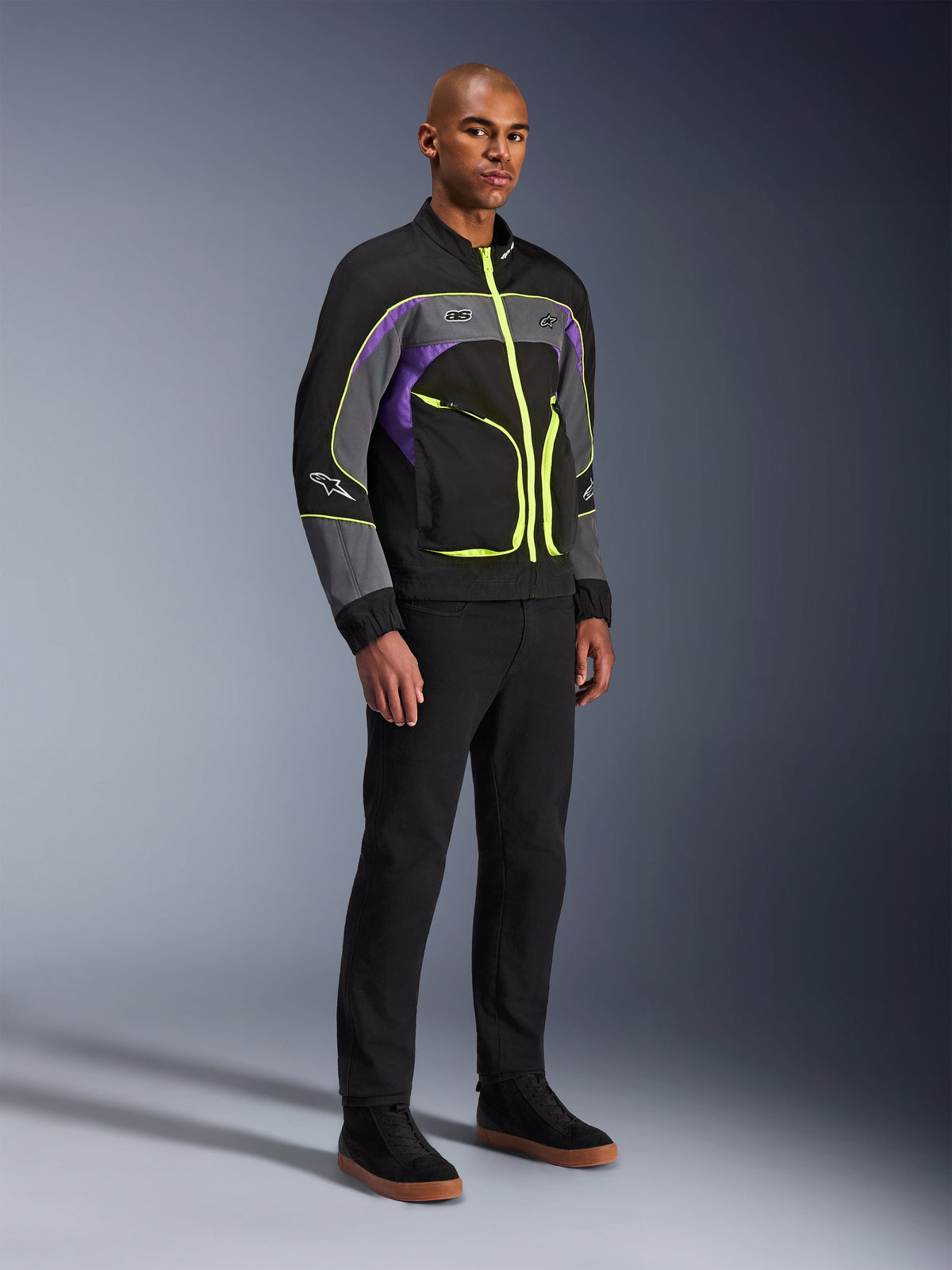Alpinestars Unite Jacket, veste de moto urbaine, Noir Asphalt Violet avec des accents jaune fluo, portée par un mannequin avec un pantalon noir et des baskets, style streetwear contemporain pour la conduite urbaine