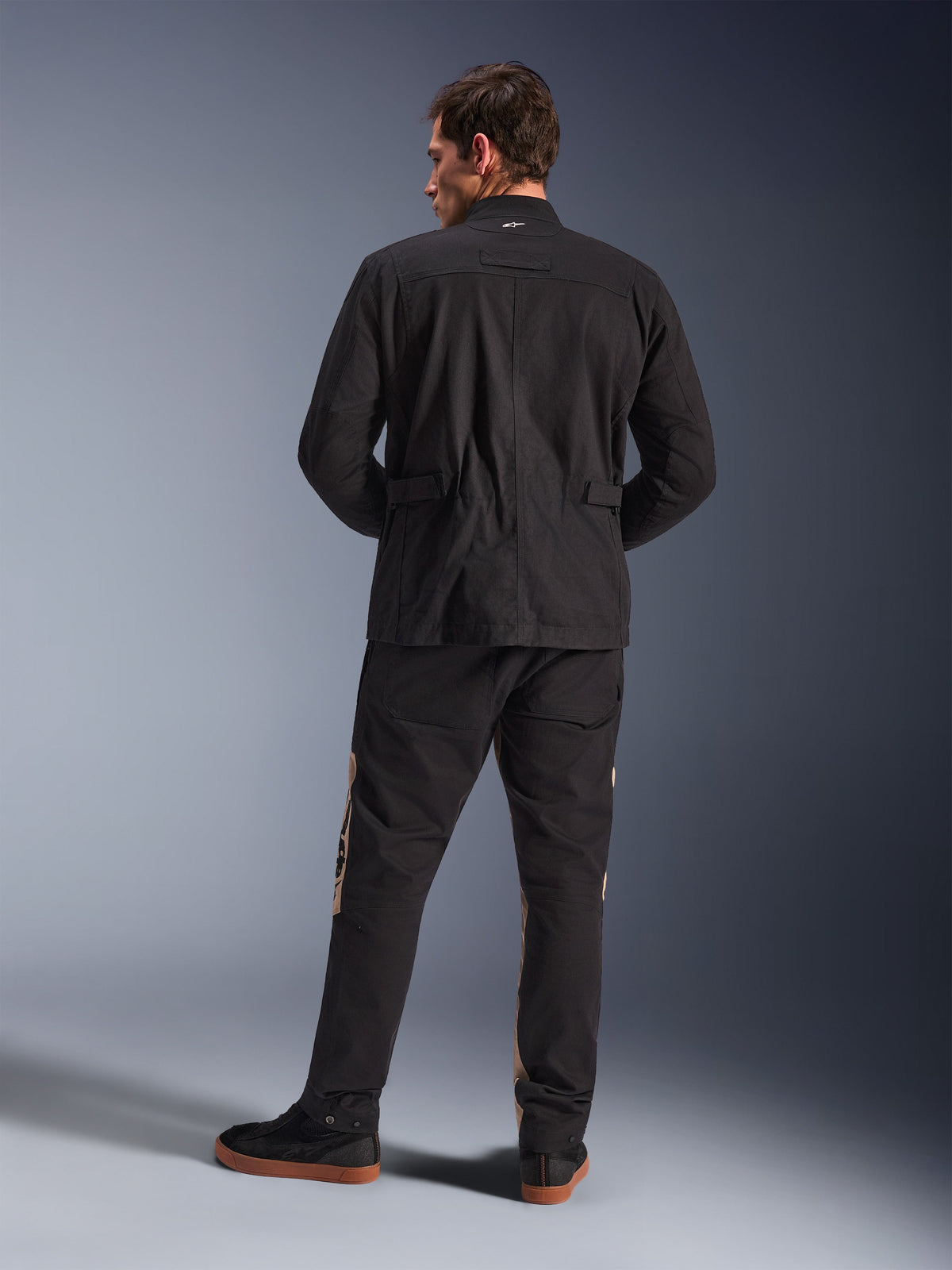Vue arrière de la veste Alpinestars Dice en noir, veste de moto urbaine avec épaules plissées et réglages à la taille, associée à un pantalon de moto noir et beige et des baskets, style urbain contemporain