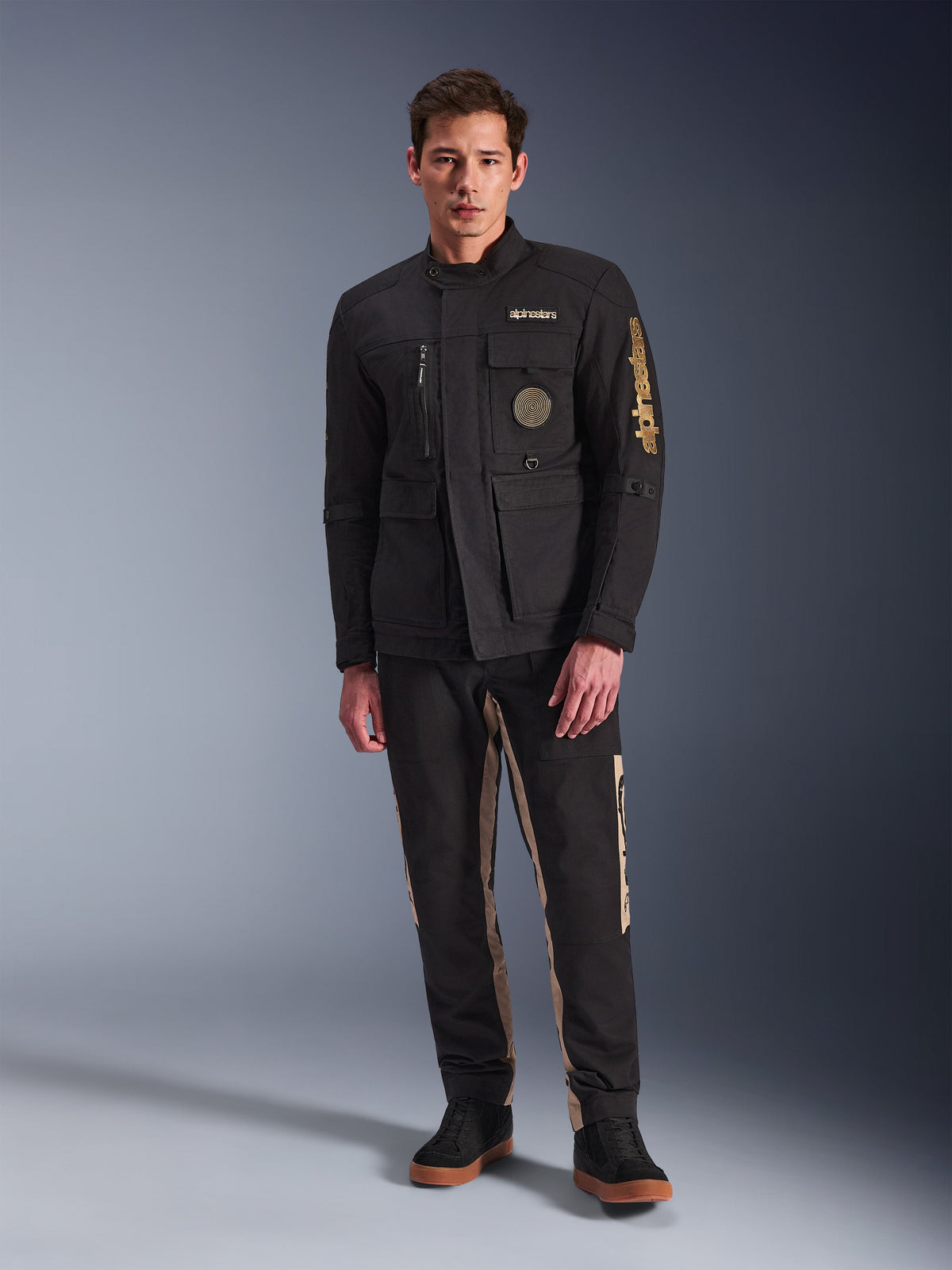 Modèle masculin portant la veste Alpinestars Dice, veste de moto urbaine noire au style field jacket vintage, quatre poches fonctionnelles à l'avant, logo Alpinestars sur la manche et la poitrine, associée à un pantalon noir et beige, équipement de conduite urbain moderne