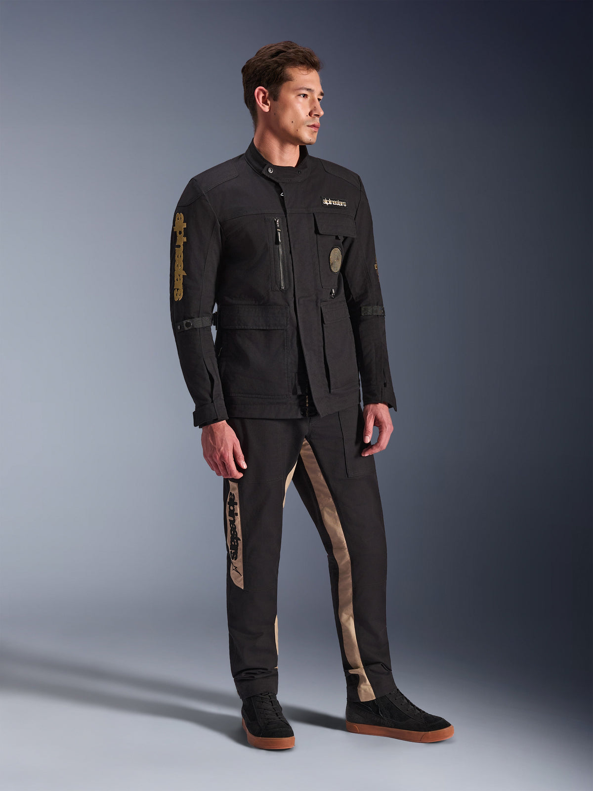 Mannequin homme portant la veste Alpinestars Dice noire, veste de moto urbaine à l'esthétique vintage field jacket, dotée de plusieurs poches utilitaires et d'un marquage doré sur la manche, avec un pantalon noir et beige assorti et des baskets noires.