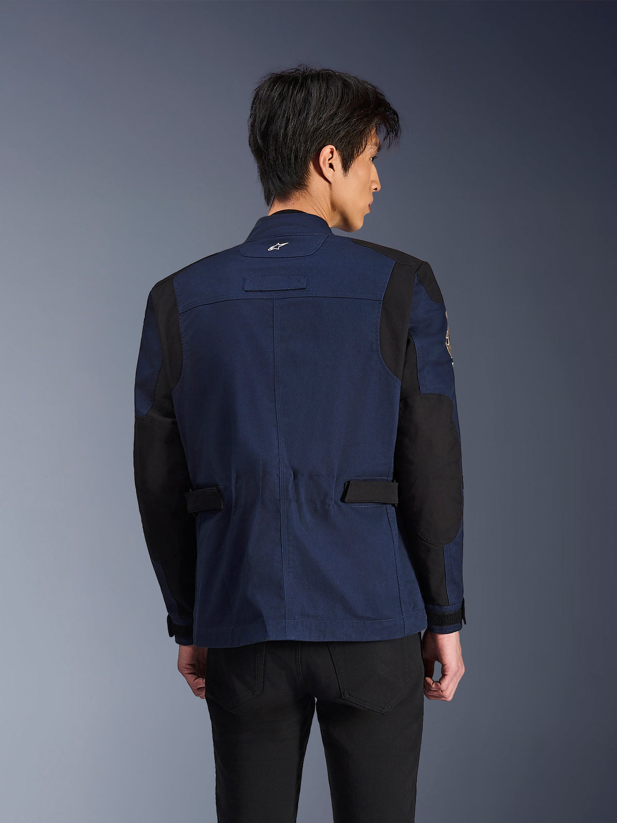 Alpinestars Dice Jacke, dunkelblaue und schwarze urbane Field-Motorradjacke, Rückansicht getragen von einem Mann, mit verstärkten schwarzen Schulter- und Ellbogenpartien und verstellbaren Taillenriegeln.