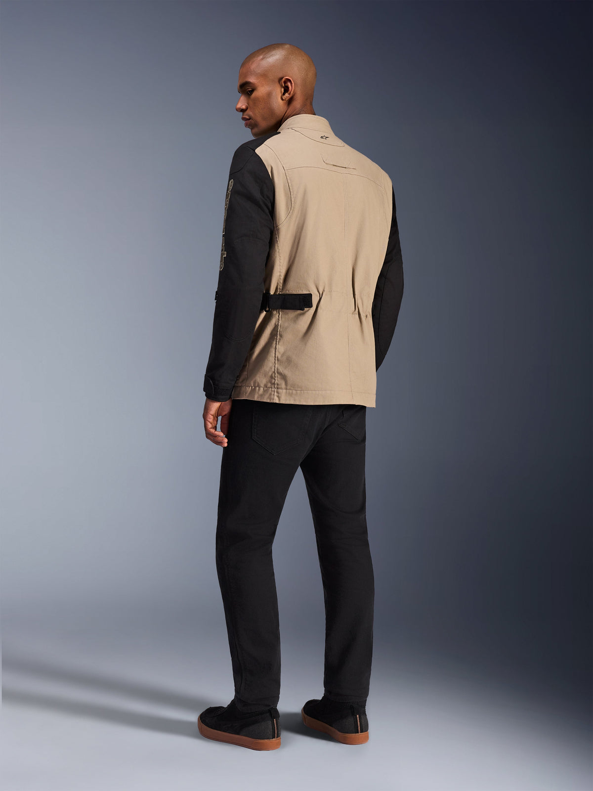 Alpinestars Dice Jacke, urbane Motorradjacke, Aluminum Black mit sandfarbenem Torso und schwarzen Ärmeln, Rückansicht am Model, robustes, von Arbeitskleidung inspiriertes Design mit Taillenverstellern, kombiniert mit einer schwarzen Motorradhose.