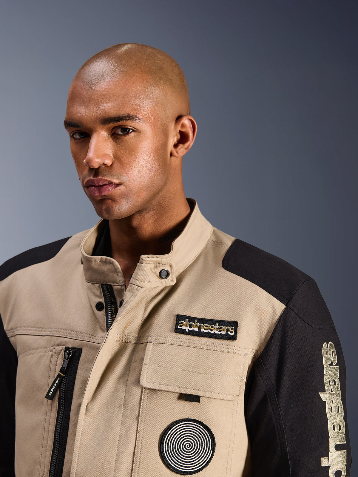 Männliches Model in der Alpinestars Dice Jacke in Aluminum Black, urbane Motorradjacke mit hellbraunen und schwarzen Farbblock-Einsätzen, bestickter Alpinestars-Brustaufnäher und Spiral-Logo, robustes, von Workwear inspiriertes Design mit hohem Kragen