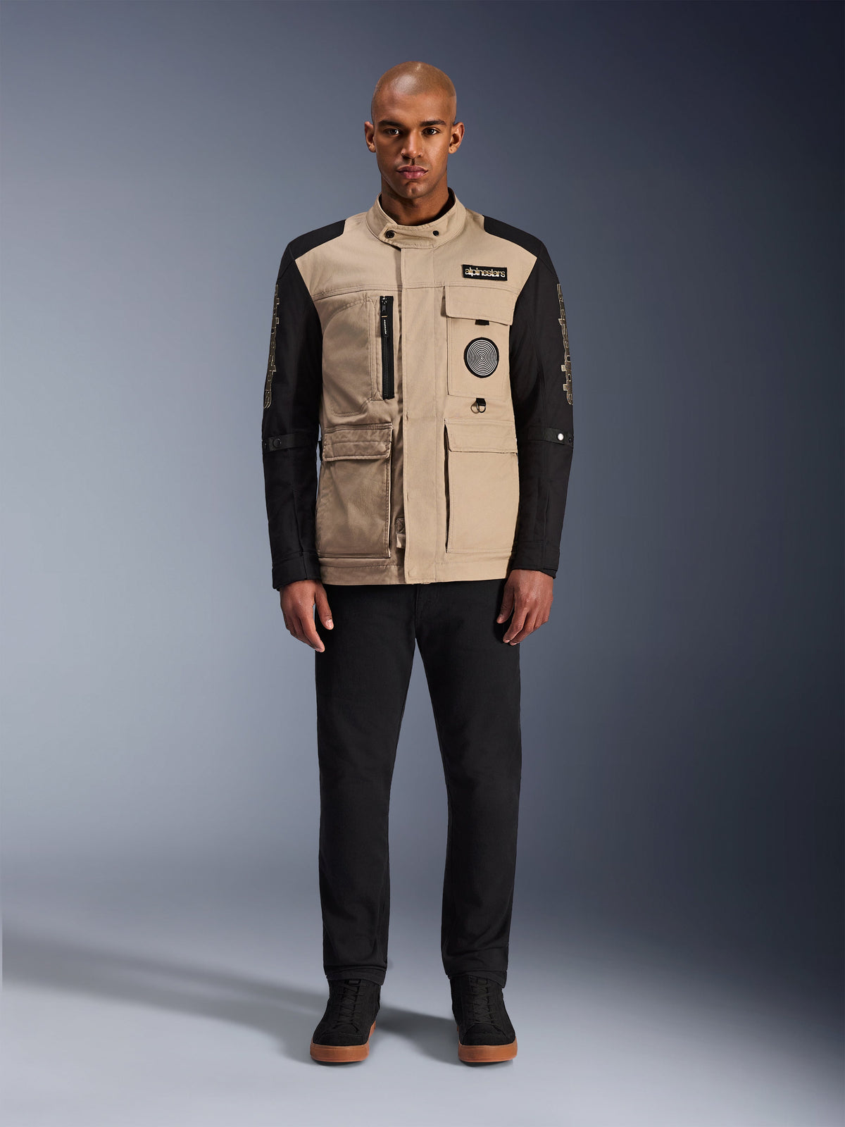 Alpinestars Dice Jacke, urbane Motorradjacke, Korpus in Aluminum-Beige mit schwarzen Ärmeln, von Arbeitskleidung inspirierte Silhouette mit vier funktionalen Fronttaschen, getragen mit schwarzer Hose und schwarzen High-Top-Sneakern, robuste straßentaugliche Fahrausrüstung