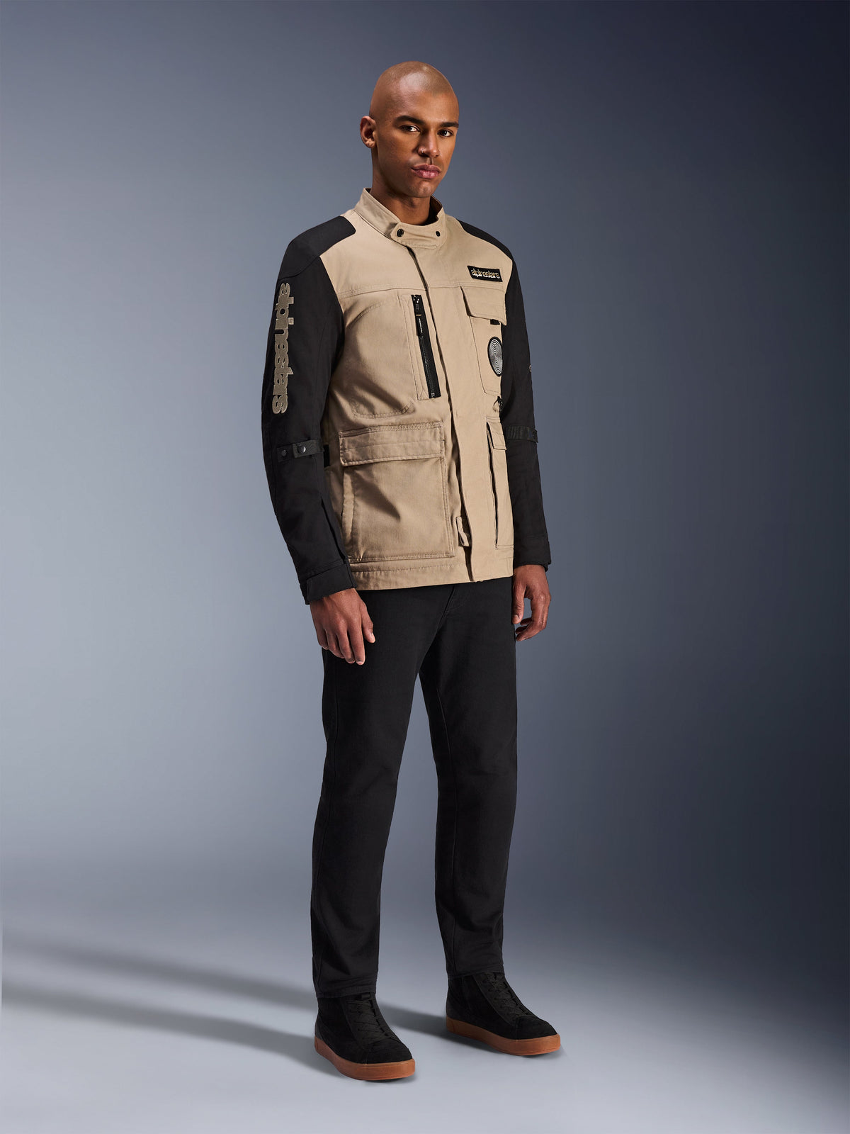 Alpinestars Dice Jacke, Urbane Motorradjacke, Aluminum Black und Tan, zweifarbiges Design mit schwarzen Ärmeln und khakifarbenem Torso, getragen von einem Model mit schwarzer Hose und schwarzen Sneakern, Workwear-inspirierte Ästhetik mit Utility-Taschen und gesticktem Branding auf dem Ärmel.