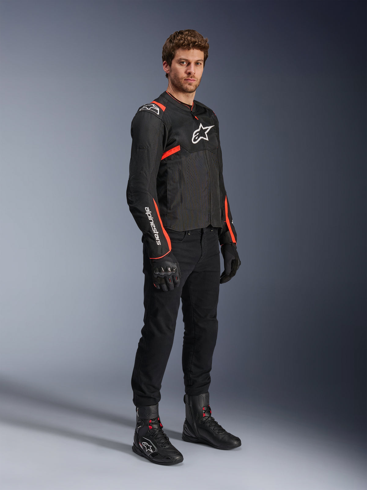 Männliches Model in der Alpinestars T-Sps Air V2 Jacke, Schwarz mit Red Fluo Akzenten, kombiniert mit schwarzer Motorradhose, schwarzen Handschuhen und schwarzen Stiefeln, sportliches Design mit Mesh-Einsätzen und weißen Alpinestars-Logos