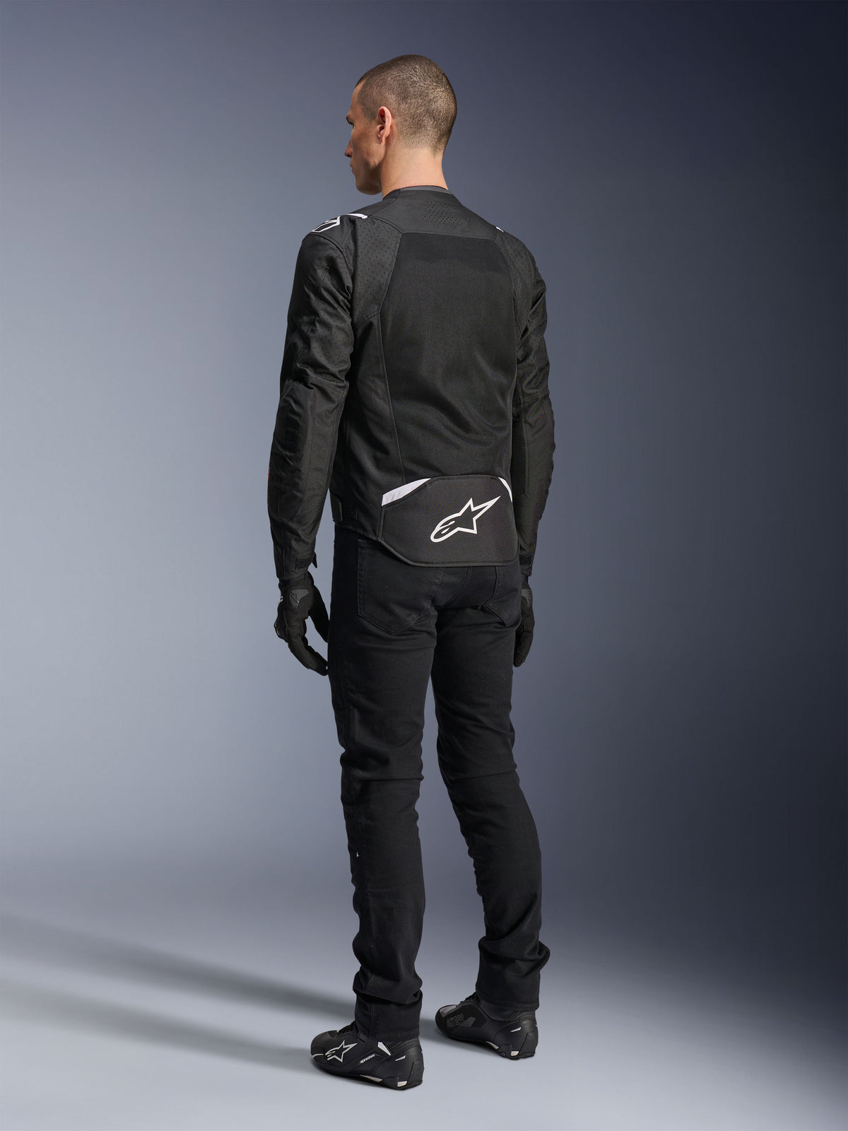 Alpinestars T-Sps Air V2 Jas, textiele motorjas, zwart, achteraanzicht met groot mesh ventilatiepaneel en wit Alpinestars-logo op de onderrug, gecombineerd met een zwarte motorbroek en handschoenen voor een sportieve urban look.