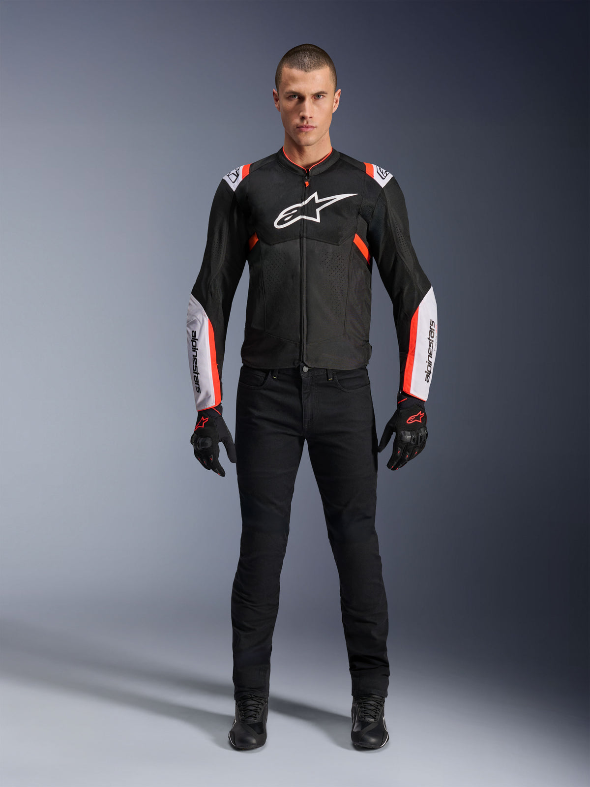 Modèle masculin portant la veste Alpinestars T-Sps Air V2 en Noir Blanc Rouge Fluo, dotée de larges panneaux en mesh technique et d'un logo blanc sur la poitrine et les manches, associée à un pantalon de moto technique et des gants noirs, équipement de moto sport urbain