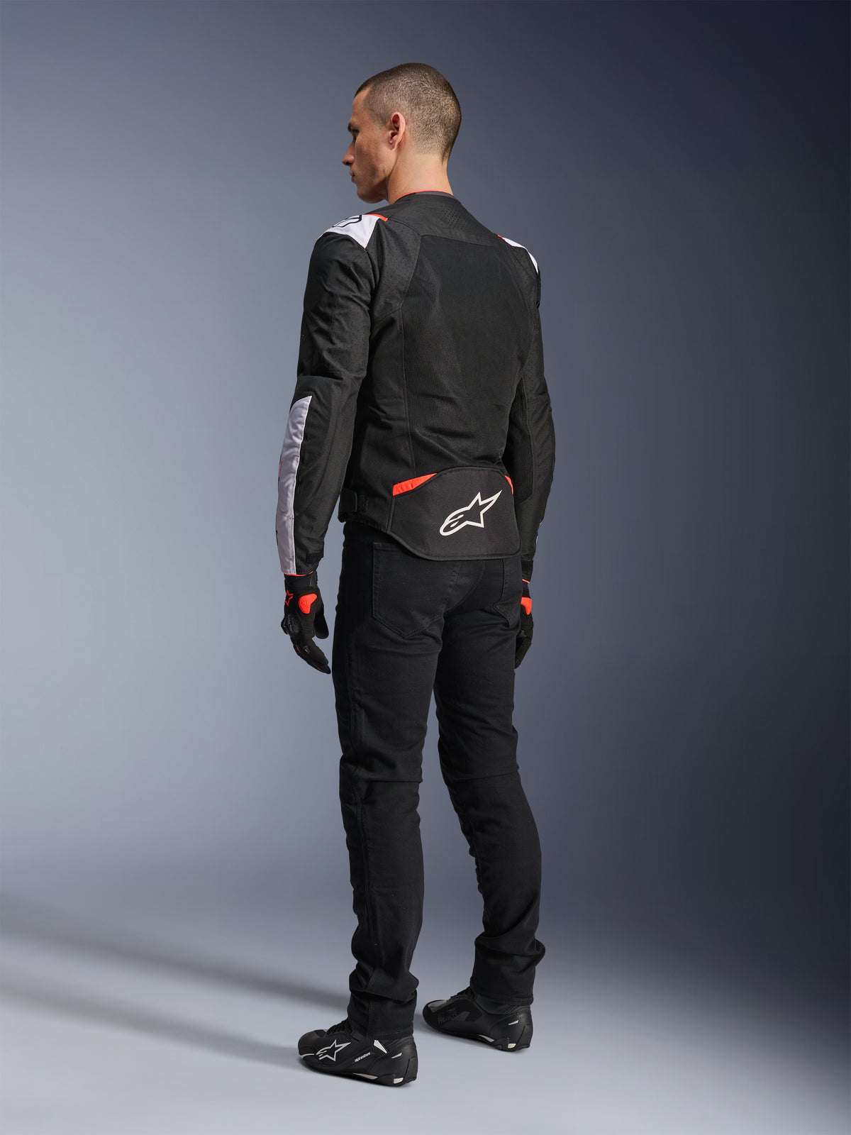 Veste Alpinestars T-Sps Air V2, veste de moto textile, noir, blanc et rouge fluo, vue arrière sur modèle, empiècements en mesh pour la ventilation, logo Astars blanc dans le bas du dos, portée avec un pantalon de moto noir et des gants