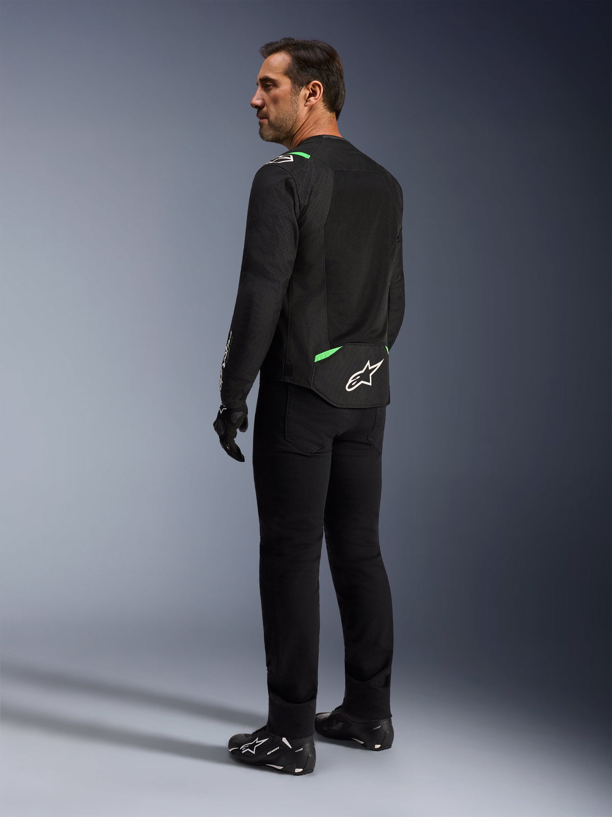 A man wearing the Alpinestars T-SPS Air V2 Jacket in Black Fluo Green, shown from a back and side profile view, styled with black riding pants and motorcycle shoes. The jacket features ventilated tech mesh panels, bright green accents on the shoulders and lower back, and a white star logo on the rear tail.