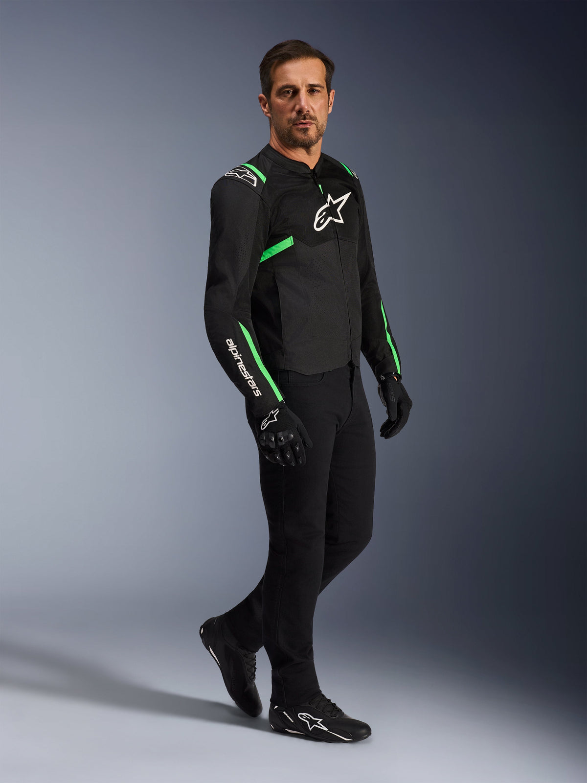 Male model wearing Alpinestars T-SPS Air V2 Jacket in Black and Fluo Green, paired with black riding pants and gloves, sport-cut textile jacket with mesh panels and neon accents, complete urban motorcycle outfit