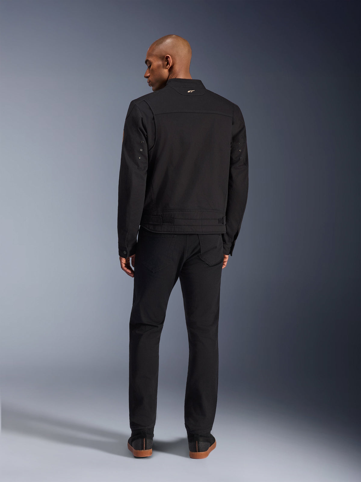 Veste Alpinestars Bruiser en noir, vue arrière d'un mannequin homme portant cette veste de moto urbaine avec un pantalon noir assorti, petit logo doré sur le col et sangles de réglage à la taille, esthétique inspirée du style workwear