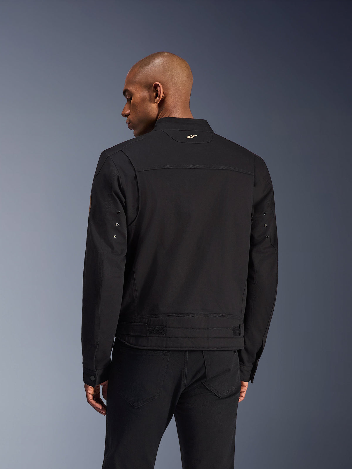 Veste Alpinestars Bruiser, veste de moto urbaine noire, vue de dos, présentant un design inspiré du style workwear avec un petit logo doré sur le col et des sangles de réglage à la taille, construction textile durable pour la conduite urbaine