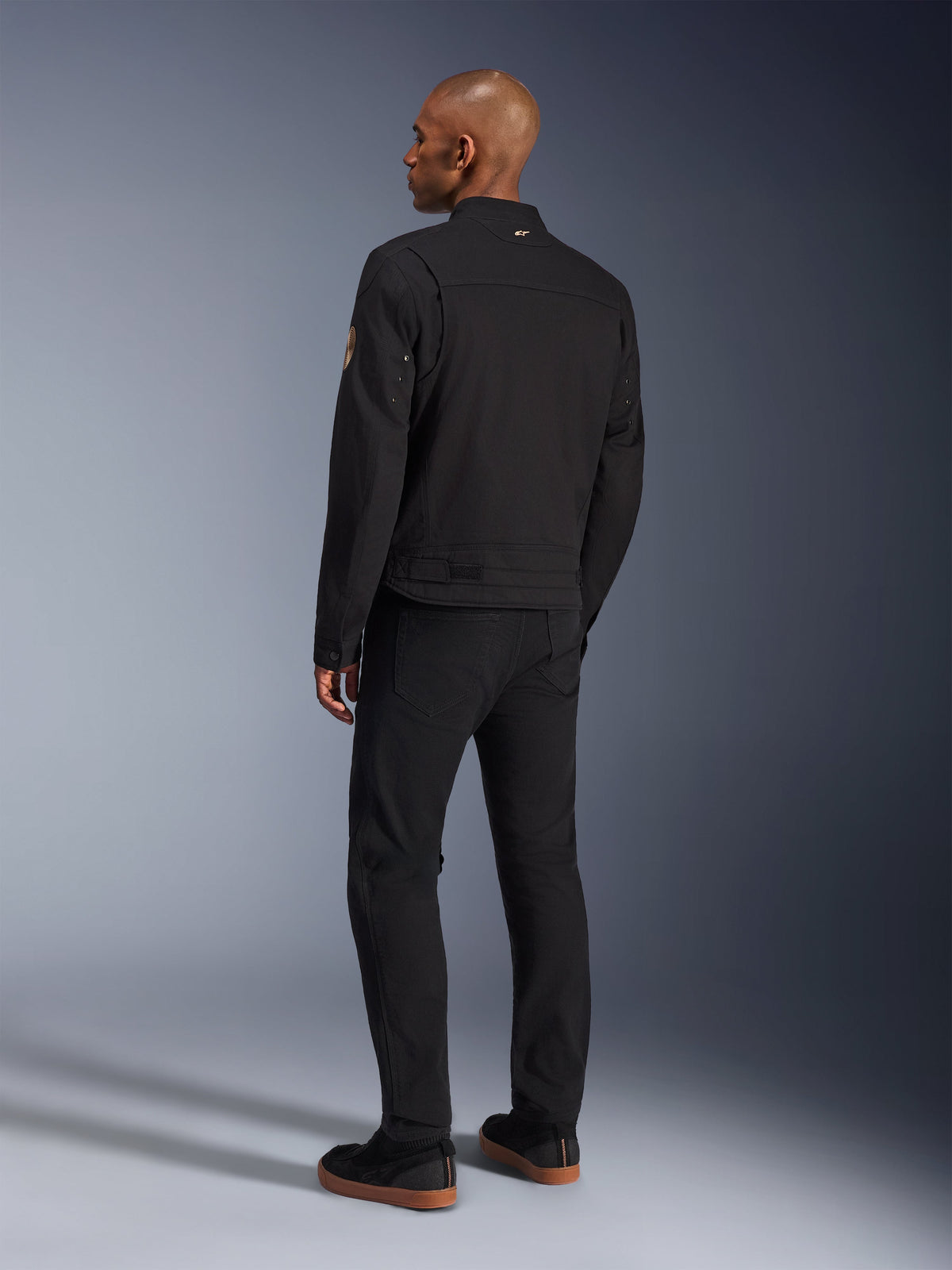 Veste Alpinestars Bruiser, veste de moto urbaine noire, vue de dos sur un mannequin, style workwear classique, avec empiècements plissés aux épaules et écusson spirale Oscar sur la manche, portée avec un pantalon noir et des chaussures noires décontractées à semelles en gomme.