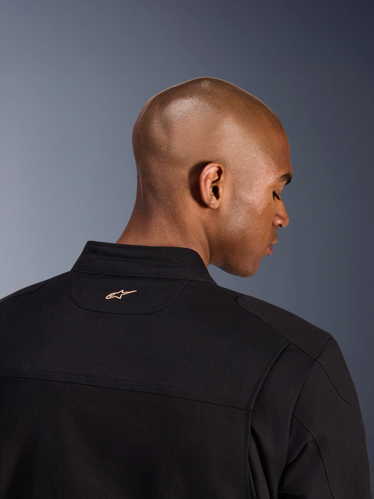 Veste Alpinestars Bruiser, noir, vue détaillée de l'arrière du col et du haut des épaules, arborant un petit logo Alpinestars brodé beige sur la nuque, veste de moto urbaine à l'esthétique workwear