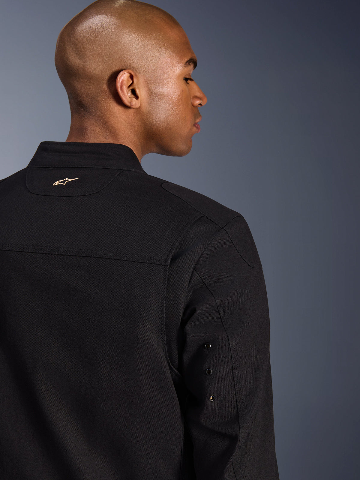 Veste Alpinestars Bruiser, veste de moto urbaine en textile, noire, vue arrière rapprochée avec logo doré brodé sur le haut du dos, détails d'œillets métalliques sur les manches et empiècements plissés aux épaules pour une mobilité accrue