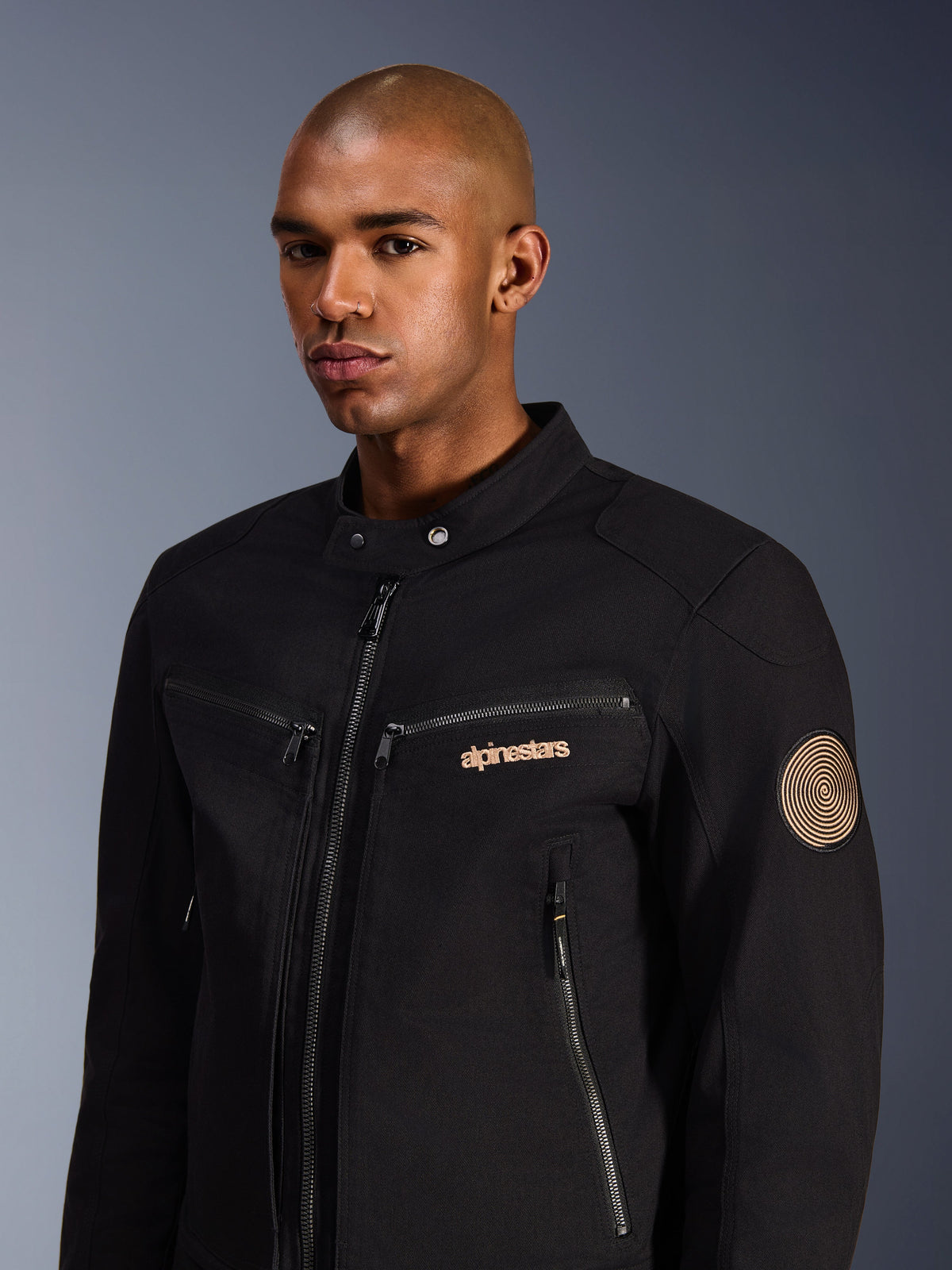 Modèle masculin portant la veste Alpinestars Bruiser, noire, veste de moto urbaine en sergé de polycoton, avec des poches zippées, un logo brodé doré sur la poitrine et un écusson en spirale Oscar sur la manche, style urbain moderne