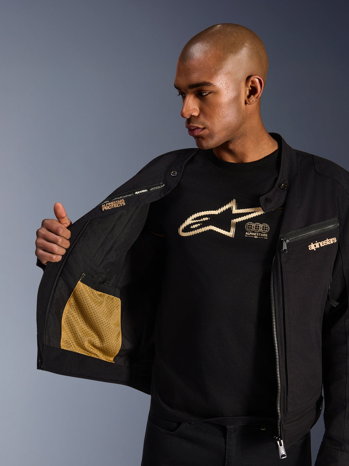 Veste Alpinestars Bruiser, veste de moto urbaine noire, vue rapprochée montrant l'intérieur ouvert avec poche en maille dorée et marquage « Alpinestars Protects », portée sur un t-shirt noir à logo, équipement de moto en textile à l'esthétique workwear
