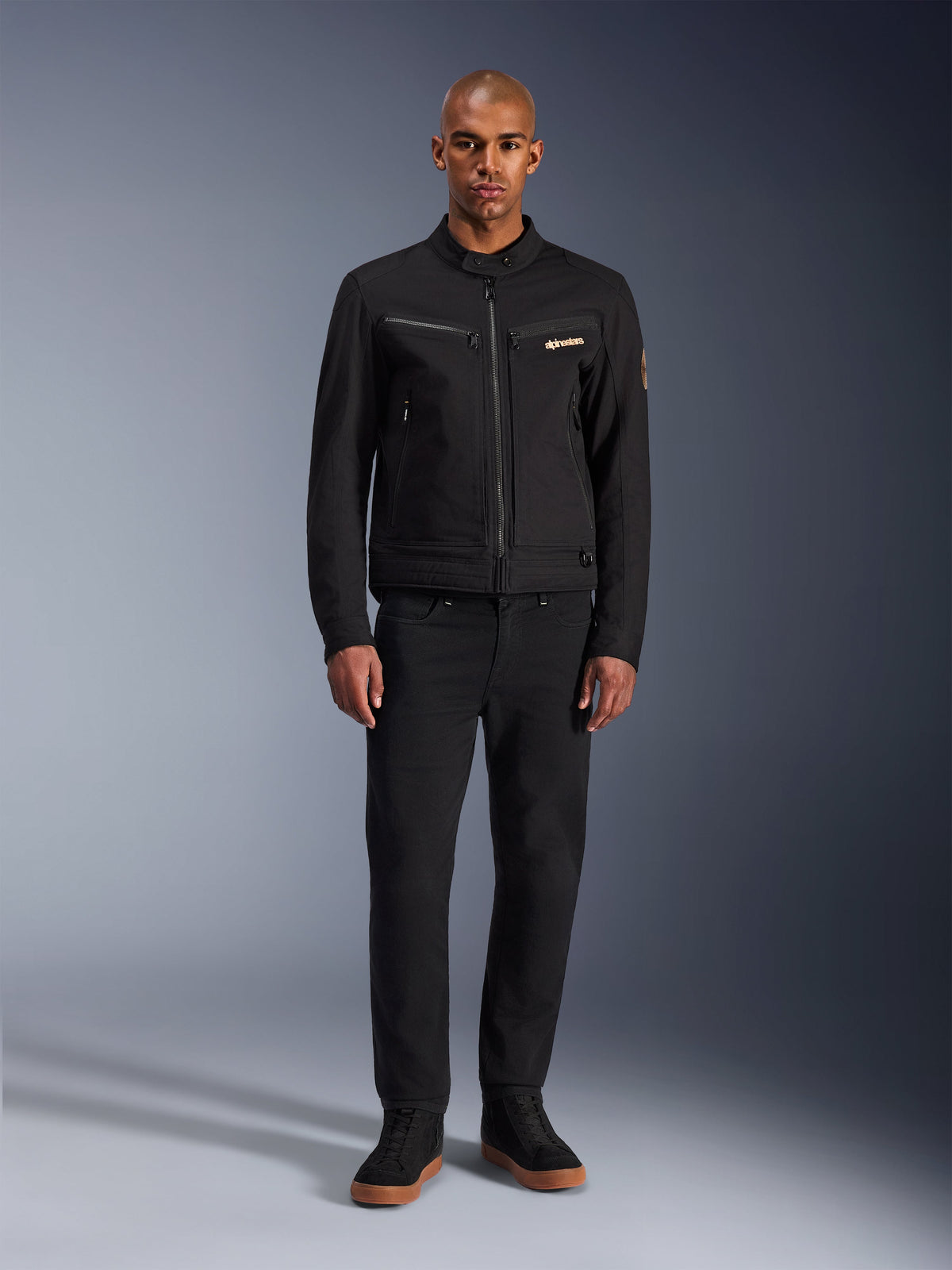 Veste Alpinestars Bruiser, veste de moto urbaine noire, portée par un mannequin avec un pantalon noir et des baskets montantes, vue de face montrant les poches de poitrine zippées et le logo brodé, design classique d'inspiration workwear pour les trajets quotidiens