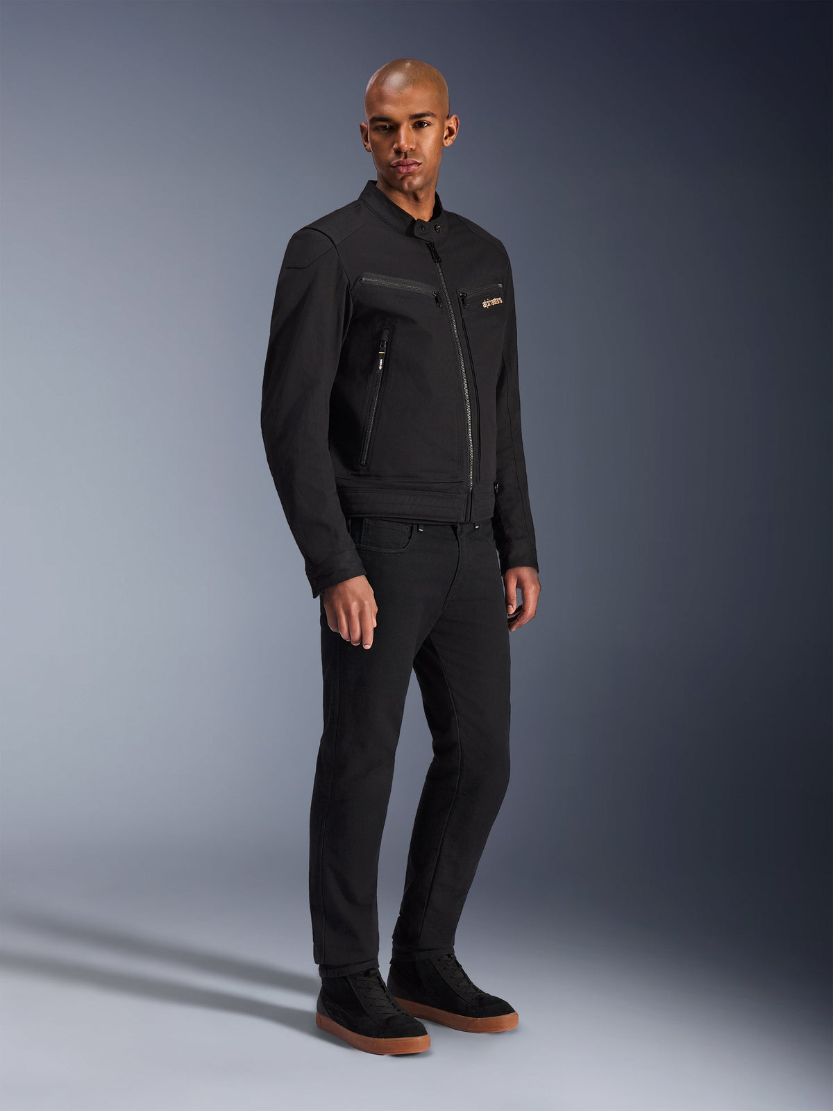 Veste Alpinestars Bruiser, veste de moto urbaine, noire, portée par un mannequin avec un pantalon et des baskets noirs, dotée de poches de poitrine zippées, d'un tissu en sergé de polycoton et d'un design élégant d'inspiration workwear pour la conduite urbaine