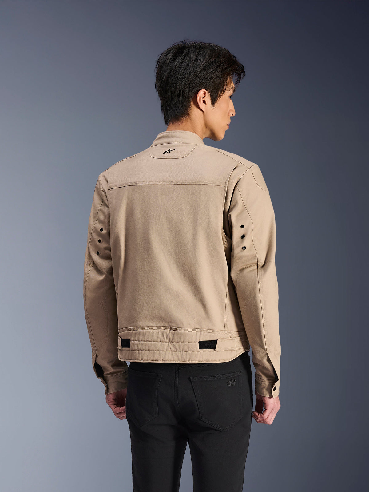 Alpinestars Bruiser Jacket, bruine urban motorjas, achteraanzicht met gearticuleerde geplooide schouders en verstelbare taillebanden, gedragen met een zwarte broek, eigentijdse op werkkleding geïnspireerde motorkleding