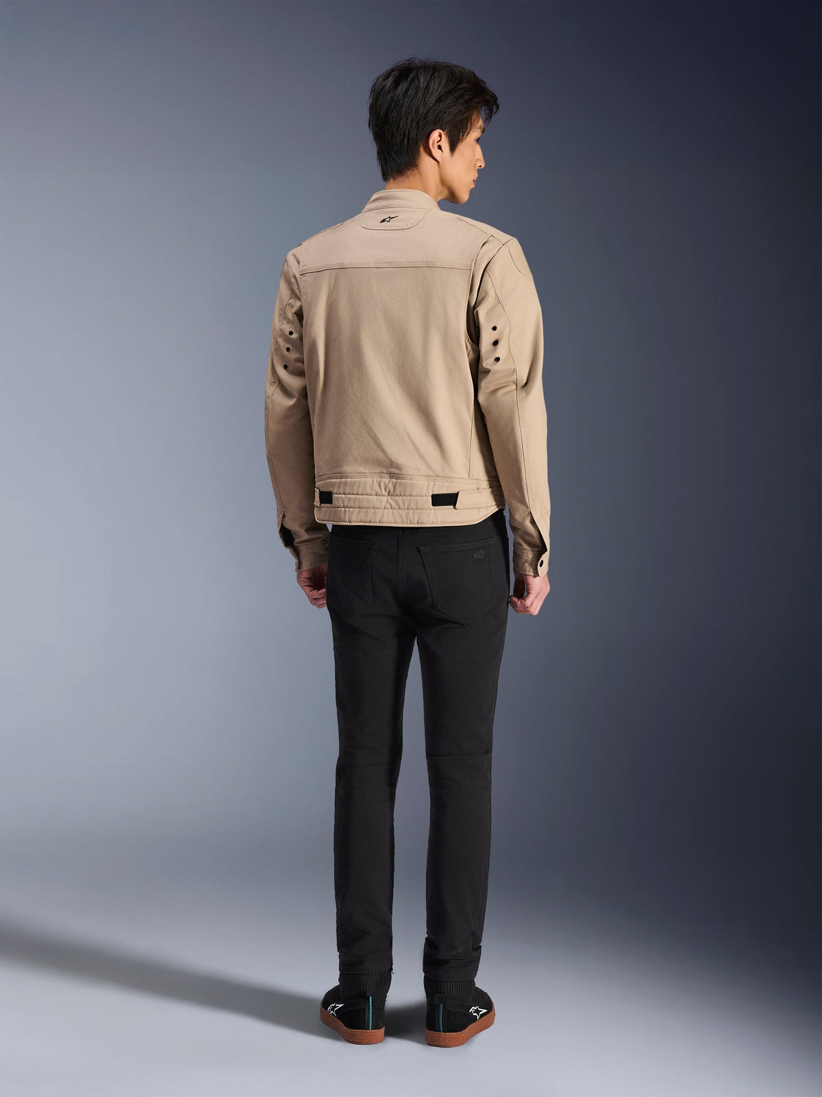 Achteraanzicht van een model dat de Alpinestars Bruiser Jacket in het bruin draagt, een stedelijke motorjas met een op werkkleding geïnspireerd ontwerp, voorzien van metalen oogjes op de rug en tailleverstellers, gecombineerd met een zwarte broek en sneakers, een eigentijdse street riding-stijl.