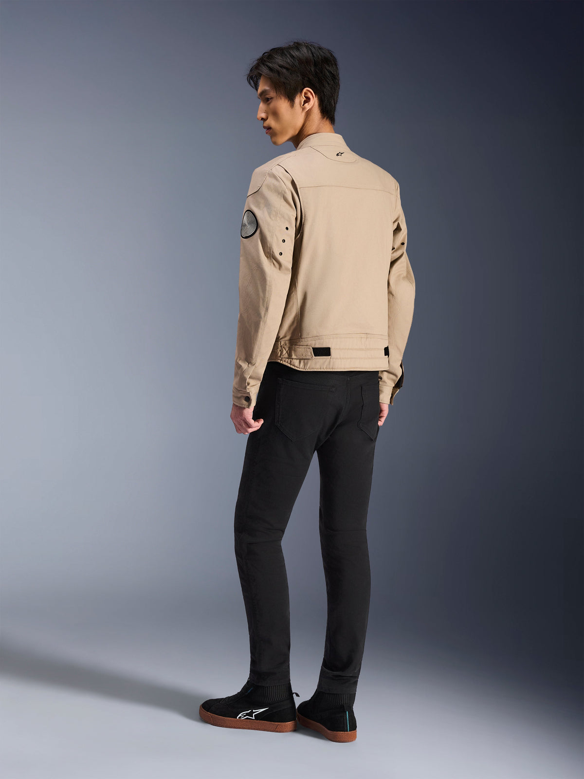 Mannelijk model van achteren die de Alpinestars Bruiser Jacket in het beige draagt, urban contemporary motorjas met werkkleding-styling, voorzien van een ronde mouwpatch, verstelbanden in de taille en gearticuleerde schouders, gecombineerd met een zwarte broek en zwarte sneakers.