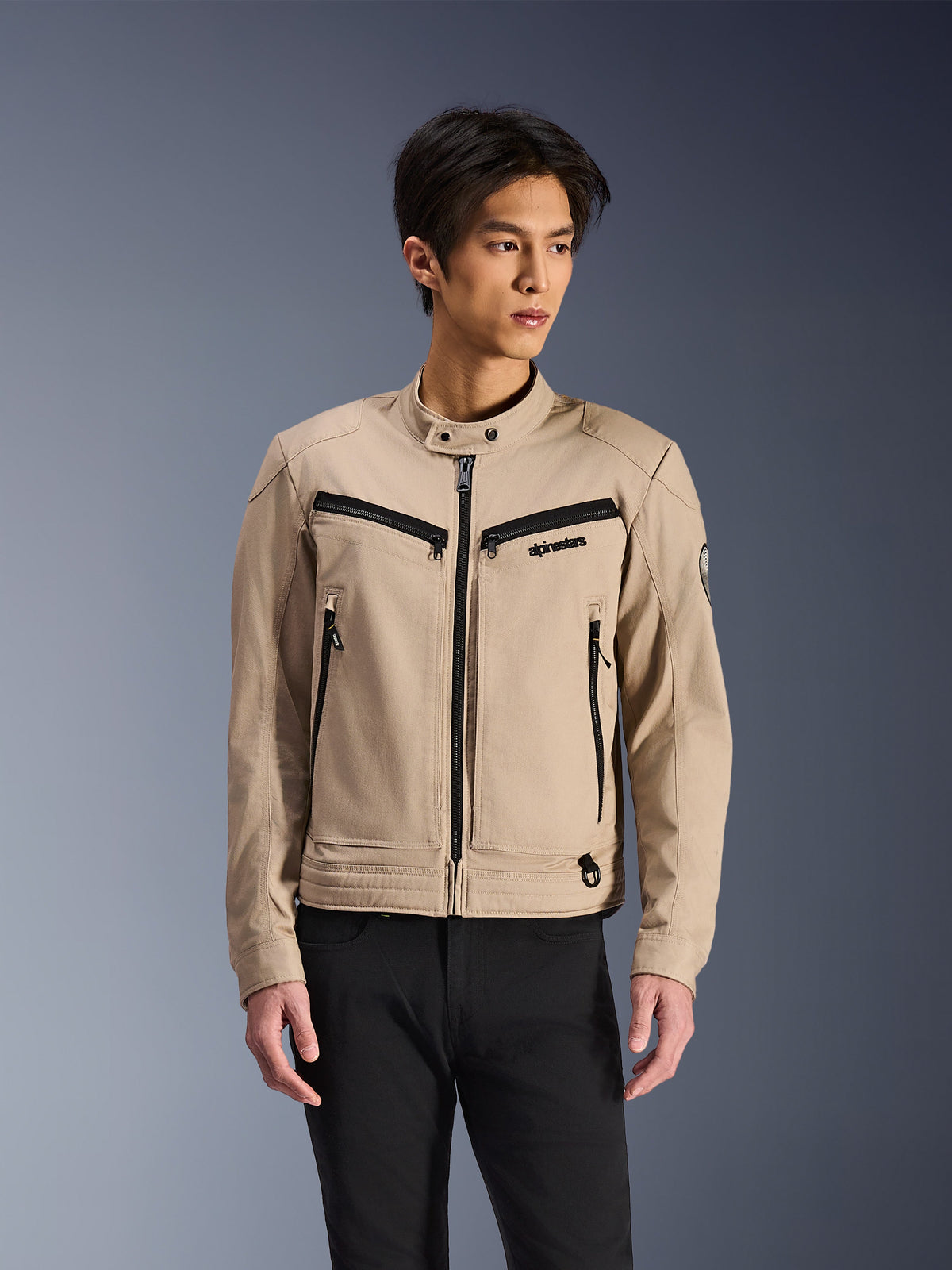 Alpinestars Bruiser Jacket, beige motorjas gedragen door een mannelijk model, voorzien van zwarte ritsdetails op de borst en zijzakken, hoge kraag met drukknopen, op werkkleding geïnspireerde urban motorkleding, gecombineerd met een zwarte broek