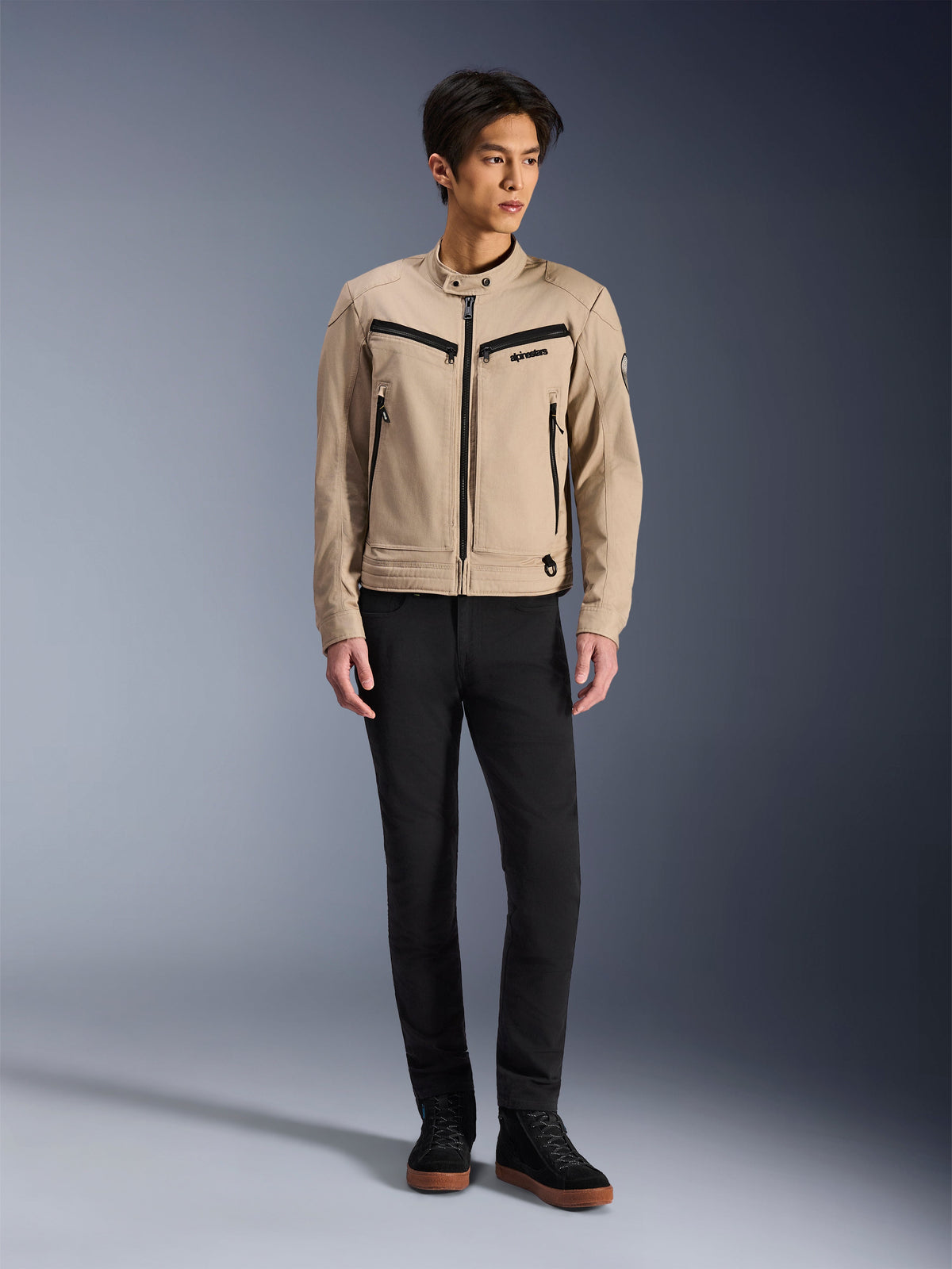 Alpinestars Bruiser Jacket, urban motorjas in de kleur Aluminum, gedragen met een zwarte broek en zwarte sneakers, voorzien van zwarte ritszakken en een kraag met druksluiting, eigentijds op werkkleding geïnspireerd ontwerp voor rijden in de stad