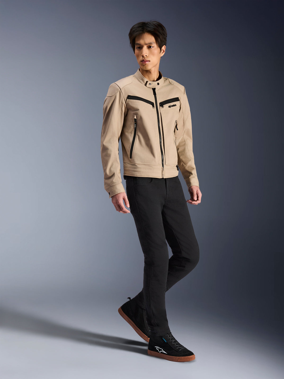 Alpinestars Bruiser jas, urban motorjas, tan, gedragen door een mannelijk model met zwarte slim-fit broek en zwarte sneakers, op werkkleding geïnspireerde textielconstructie met zwarte ritsaccenten, eigentijdse stedelijke streetstyle.