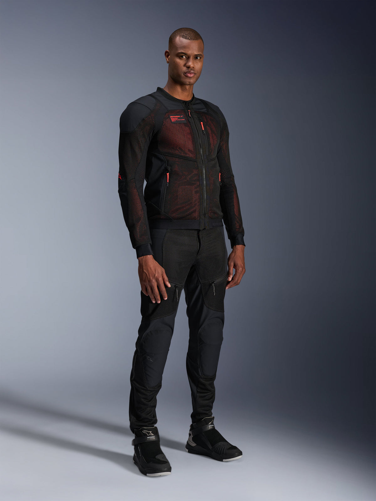 Alpinestars Troop-Air Pro Jas in Black Red Fluo, technische gepantserde mesh motorjas gedragen door een mannelijk model met bijpassende zwarte gepantserde broek en hoge motorlaarzen, volledige studio-opname.