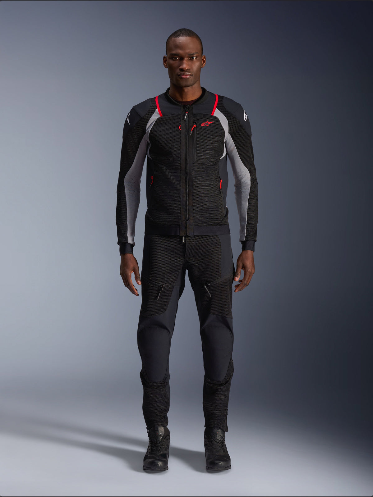 Veste Alpinestars Troop-Air Pro, veste de moto technique, noir, gris cendré et rouge vif, avec panneaux en mesh et accents rouges, portée par un mannequin homme avec un pantalon de moto et des bottes noirs, design urbain épuré avec ventilation technique.