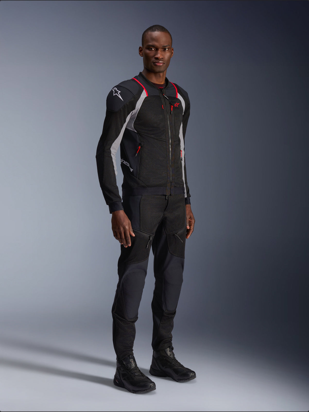 Veste Alpinestars Troop-Air Pro, noir, gris cendre et rouge vif, veste blindée technique en mesh, avec accents rouges et logo blanc sur l'épaule, portée avec pantalon et bottes noirs assortis, équipement de moto urbain moderne