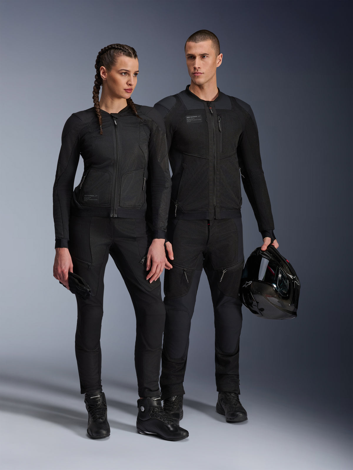 Veste Alpinestars Stella Troop-Air, homme et femme portant un équipement de moto technique assorti tout en noir, doté d'une construction en maille pour la circulation de l'air, pantalons et bottes noirs, homme tenant un casque noir, tenue de moto urbaine