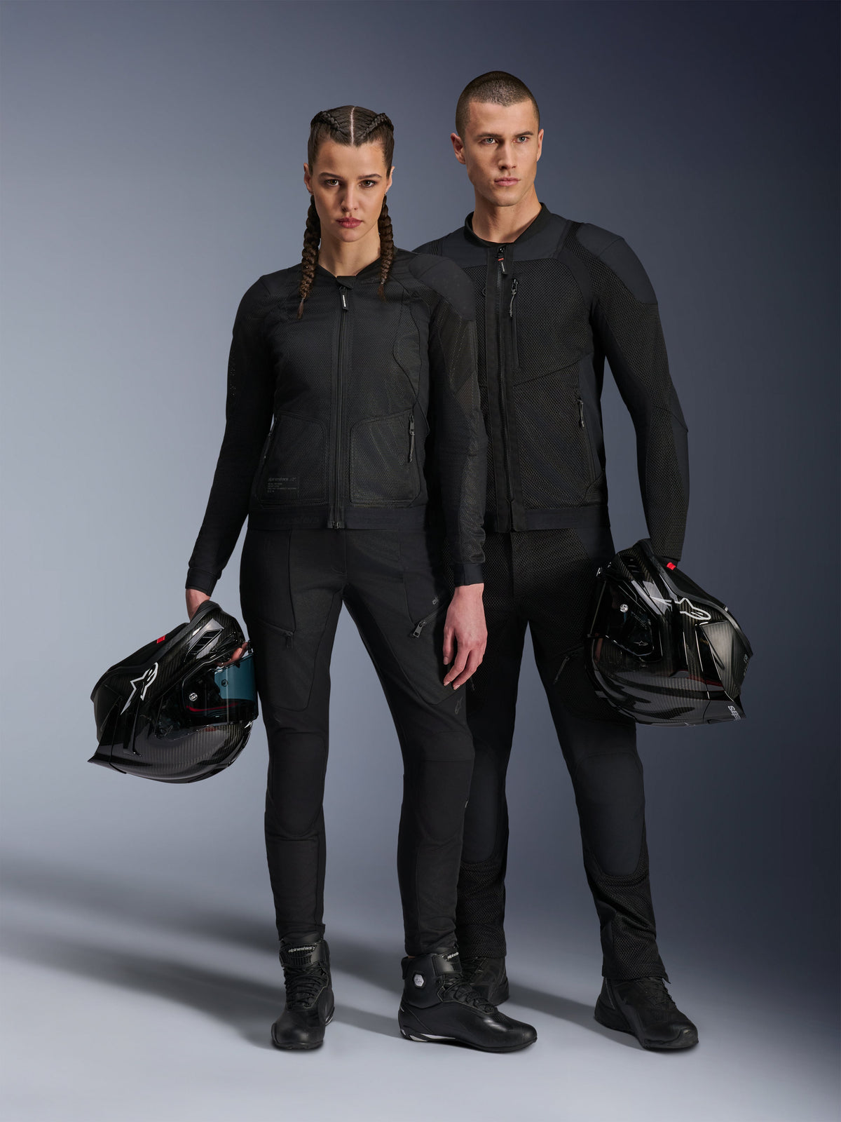 Männliches und weibliches Modell tragen die Alpinestars Troop-Air Pro Jacken in Schwarz, technische Mesh-Fahrerausrüstung mit Protektoren kombiniert mit passenden schwarzen Hosen und Stiefeln, beide halten einen schwarzen Helm und präsentieren koordinierte urbane Motorradbekleidung.